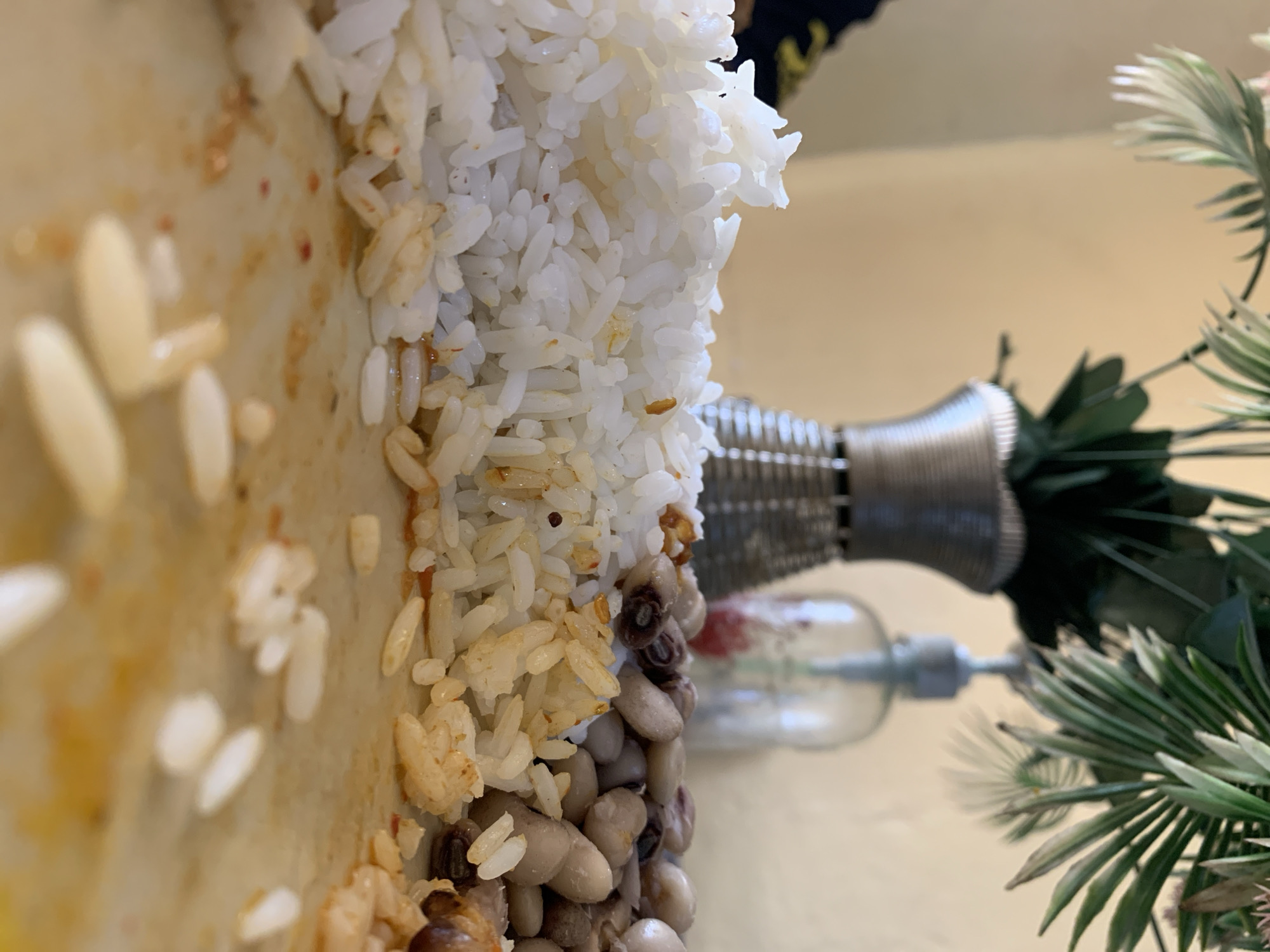 The image presents a low-angle, close-up perspective of a meal on a white surface, consistent with a plate, situated in New Stadium, Nigeria. The foreground displays remnants of food, including a pile of white rice on the left, an adjacent portion of yellowish-brown rice, and a collection of light-colored cooked beans (likely black-eyed peas) to the right. Loose grains of rice and reddish-brown food stains are scattered across the plate surface. In the blurred midground, a beige-toned wall is visible. A dark-colored fabric with a yellow pattern, partially visible on the far left, indicates the presence of an individual. A silver-toned, horizontally-ribbed vase containing artificial green, palm-like foliage stands centrally in the midground. To its right, a clear glass dispenser bottle with a grey pump top is also present, out of focus. The background consists of the continuous beige wall.