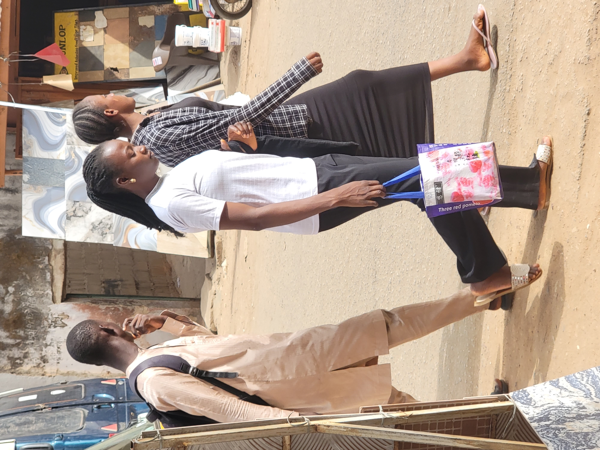 In the urban environment of Maiburiji, Nigeria, three individuals are captured on what appears to be a street or sidewalk. One man, wearing a light brown shirt and black backpack, stands near what seems to be a metal structure and tiled merchandise. He has dark skin and short hair. Two women are walking together next to him. The woman closer to the camera is wearing a white shirt and black pants, carrying a shopping bag. The second woman is wearing a black and white checkered shirt and a long black skirt. Both women have dark hair, with the first woman's hair braided long. The environment consists of a paved surface and various buildings in the background, including one with tiles and a yellow sign. The scene is brightly lit.