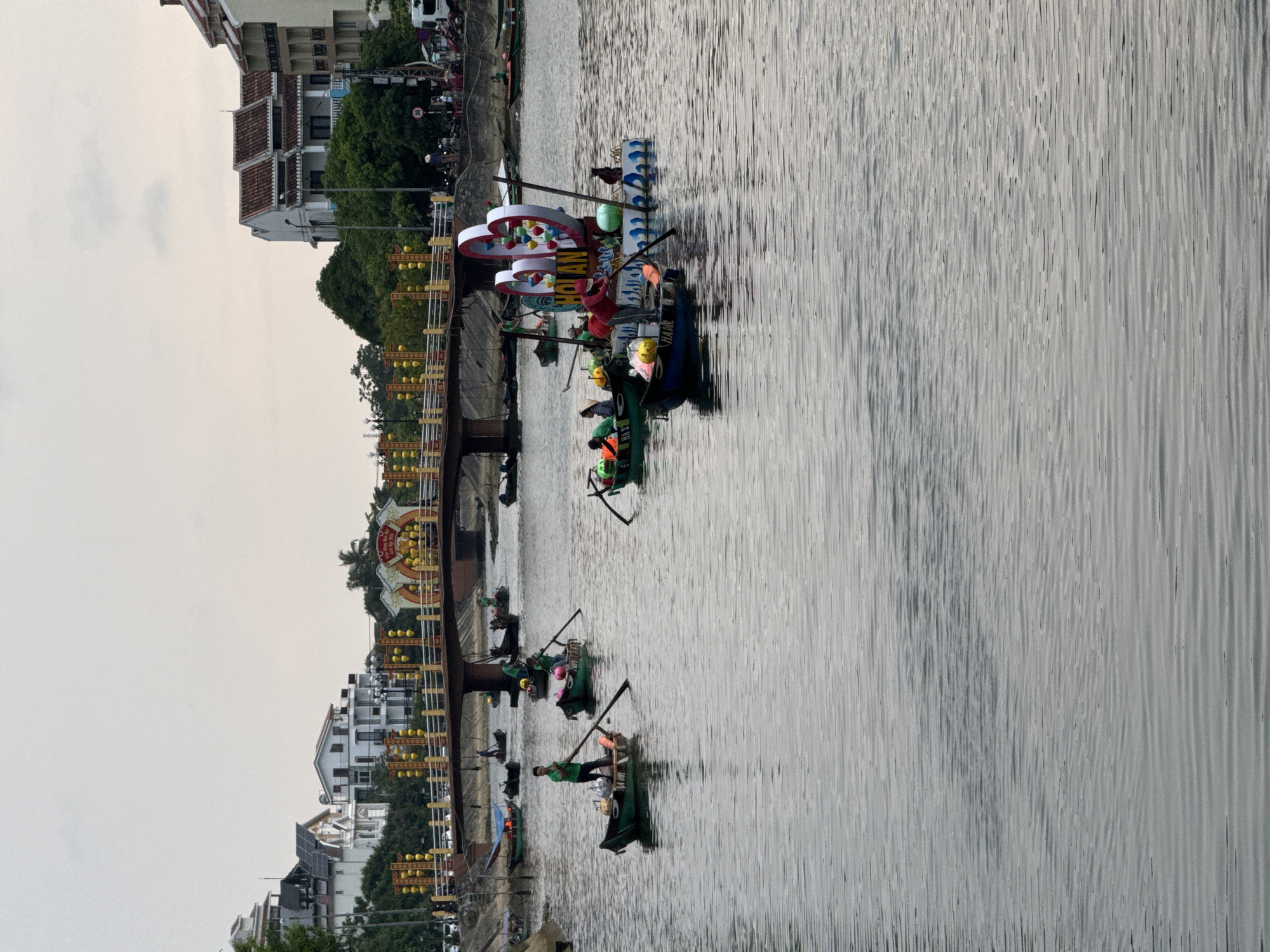 This image captures a lively scene on a river in Hội An, Vietnam, under an overcast daytime sky. The river's surface shows subtle ripples, reflecting the soft light.
Numerous small, traditional Vietnamese boats, likely sampans, are scattered across the water. Several green-colored boats carry one or two individuals, many of whom are wearing yellow or green life jackets. These people are actively paddling with long oars, some standing, some seated, suggesting a recreational or tourist activity such as a boat tour.
A larger, more ornate boat stands out with elaborate decorations, including prominent red and white heart-shaped structures adorned with colorful lanterns. On this boat, the text "HỘI AN" and "HAW" is clearly visible on a banner. Another smaller green boat has partial text visible, possibly "NHA..." or "HHA...".
The riverbanks are lined with buildings exhibiting traditional Vietnamese architecture, characterized by tiled roofs and multi-story structures, interspersed with lush green trees. A significant feature is a bridge, heavily decorated with numerous yellow lanterns and banners, which are characteristic of Hội An's charming aesthetic. The overall scene suggests a vibrant cultural experience, with tourists or locals enjoying the tranquil river environment and the festive decorations of the historic town.