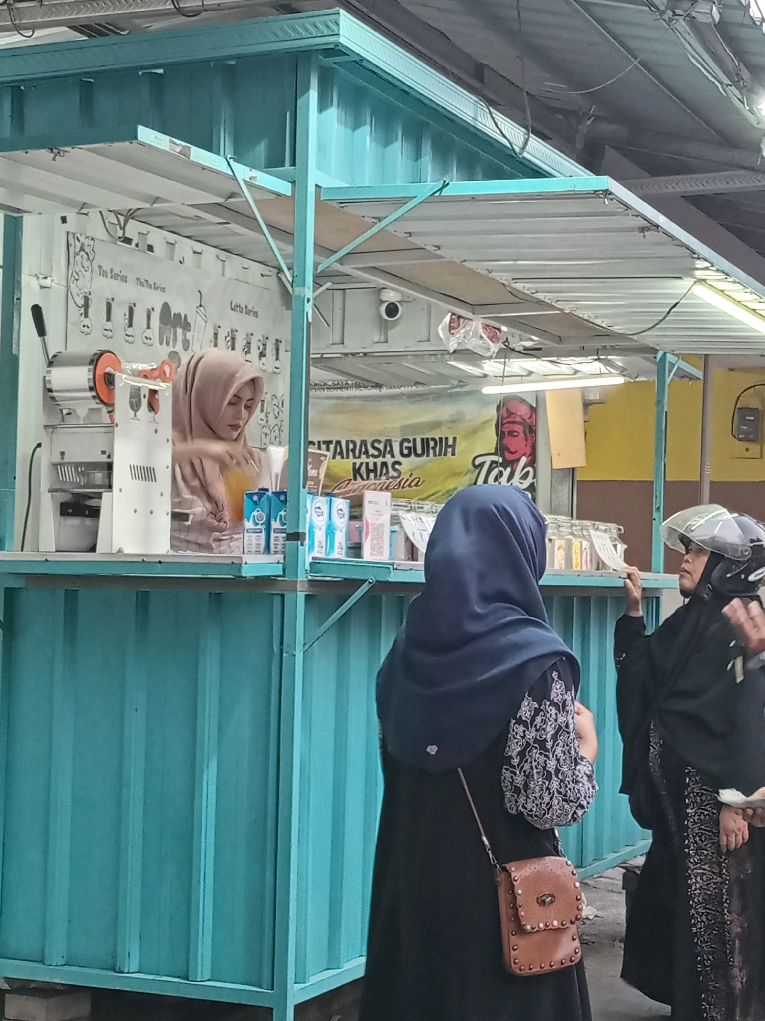 In Randuagung, Indonesia, a food stall with a turquoise corrugated metal facade and a white corrugated metal awning is depicted. A woman wearing a light brown hijab is behind the counter, preparing a beverage in a clear cup with a yellow liquid. To her right are several small, blue and white cartons. In front of the stall, two women stand. The woman on the left wears a dark blue hijab and a black abaya with white embroidered sleeves, carrying a small brown leather purse. The woman on the right wears a black hijab and abaya, and a helmet, appearing to be conversing with someone to her right. A banner behind the counter features text in Indonesian and a graphic of a man's head. Various items, including a drink dispenser and packaging, are visible on the stall's shelves. The environment appears to be an outdoor marketplace.