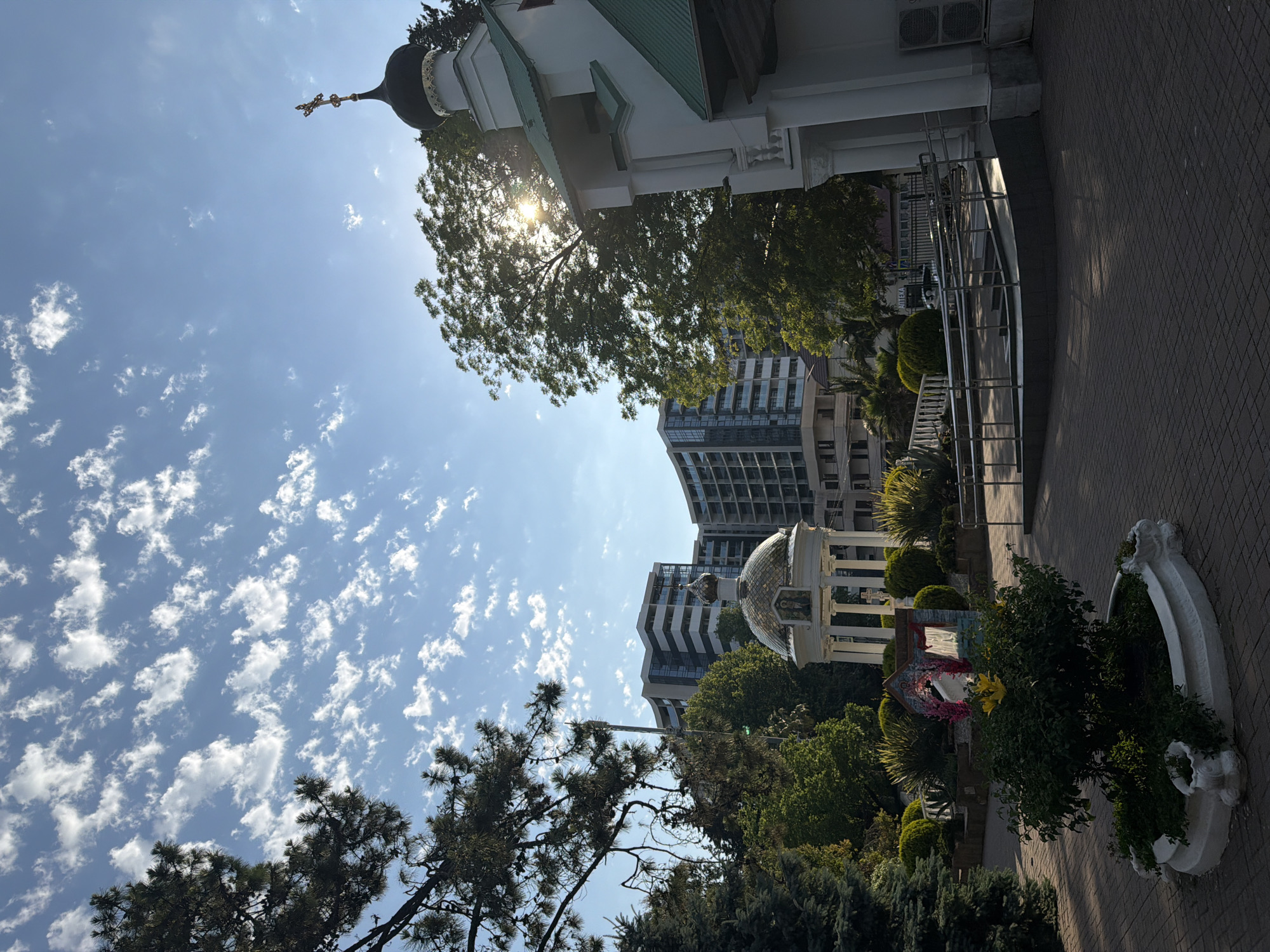 This media file depicts an outdoor scene under a bright, sunny sky with scattered white clouds. The sun is visible, shining through the branches of a mature evergreen tree, casting sunbeams. In the background, several modern apartment buildings rise behind lush green trees and ornamental shrubs. A white building with a green roof and a black dome topped with a cross is visible on the left, suggesting it might be a church or a similar religious structure. A paved walkway with a metal railing runs along the right side of the frame. Near the walkway, there is a decorative white planter filled with greenery, and further back, a small ornate structure with a golden dome. The overall impression is of a warm, clear day in a semi-urban or resort-like setting, possibly in Sochi, Russia, given the context. There is no visible activity of people. The time of day appears to be late morning or early afternoon due to the high angle of the sun.