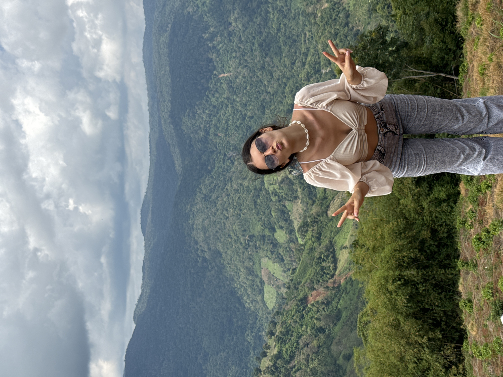 A young woman with dark hair, wearing a beige crop top, grey jeans, and sunglasses, poses for a photo with a scenic mountain landscape in the background. The woman is making peace signs with both hands. The mountainous terrain is covered in dense green vegetation, with some patches of cultivated land visible on the slopes. The sky is overcast with dramatic clouds. The image appears to be taken outdoors in a natural setting. The location is identified as Nao Se, Vietnam.