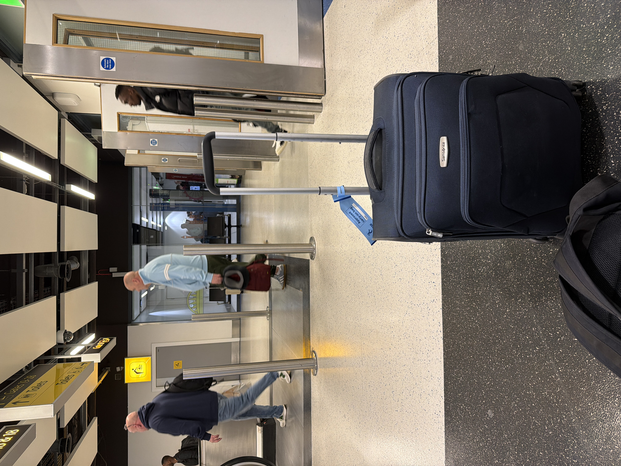 The image depicts a scene inside Gatwick Airport, located in Crawley, United Kingdom. A blue Samsonite carry-on suitcase with an "Approved Carry-on Bag" tag on the handle is the central object, positioned on a floor with contrasting white and black speckled tiles. Passengers are queuing and walking through security lines. A man in a blue jacket and jeans walks past the metal barriers, while another man in a light blue shirt stands in line. An additional person in a black jacket is seen entering a passageway marked with a blue sign. Overhead signs indicating directions to gates and toilets are visible on the left side of the frame. Fluorescent lights illuminate the area.