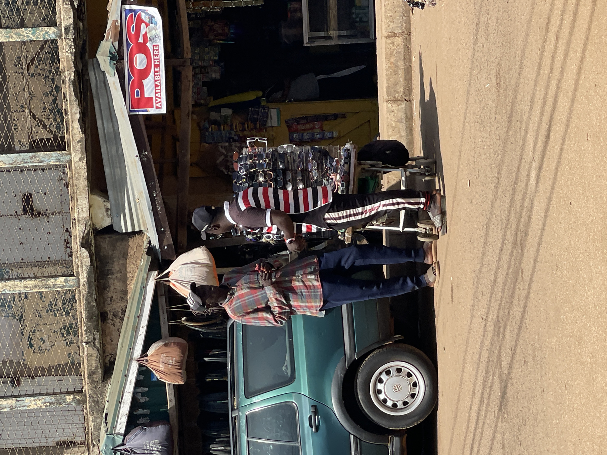 The image depicts a street scene in Bukuru, Nigeria. Two men are standing in front of a green Volkswagen van parked beside a storefront. The man closer to the viewer is wearing a plaid shirt and jeans. The other man is wearing a black hat, a striped shirt, and pants with white stripes down the side. The storefront displays various items for sale, including sunglasses. A sign above the store indicates "POS AVAILABLE HERE". The background reveals other stores and goods. The street itself appears to be paved and moderately lit.