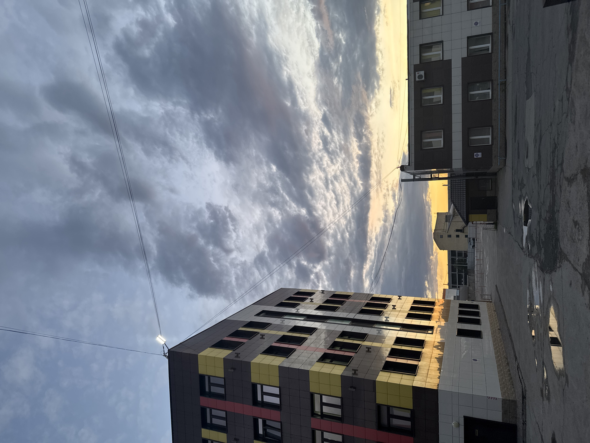 The image, rotated ninety degrees counter-clockwise from its intended orientation, captures an urban scene in Perm, Russia, at either sunrise or sunset. Two multi-story buildings frame a dramatic sky filled with textured clouds.
The prominent building on the left (when correctly oriented) features a modern facade with dark gray, yellow, and red paneling, punctuated by numerous windows. A bright outdoor light fixture is visible on its upper corner, from which thin power lines extend across the cloudy sky. The building on the right is characterized by white and dark brown paneling, rectangular windows, and an air conditioning unit mounted on its exterior.
The ground between the buildings appears to be an asphalt or concrete surface, marked with cracks and several puddles. These puddles reflect the sky and the glowing light from the horizon. The sky itself is a notable feature, displaying heavy, dark clouds that gradually give way to a luminous golden light on the right, indicating the time of day. This warm, golden light reflects off the windows of the left building and the water puddles on the ground.
No people or specific activities are visible in the static scene. There is no discernible text or branding visible on any of the buildings or in the immediate surroundings.