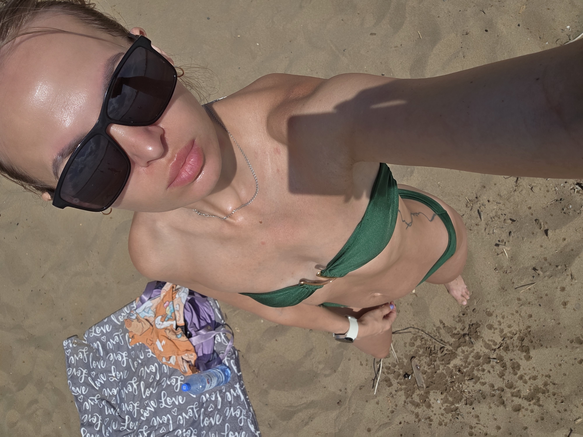A high-angle medium shot shows a woman in a green bikini standing on a sandy beach. She is wearing large black sunglasses and a silver necklace. Her body is tan, and she appears to be applying something to her lips. To her left, a patterned blanket with the word "love" repeated in cursive is spread on the sand, with a bottle of water and some clothing on top of it. The woman's right arm is bent at the elbow, and she is wearing a white watch. The sun is shining, casting a shadow on the sand from the woman's arm. The overall impression is that of a sunny day at the beach. The visible text on the blanket reads "Love" multiple times.
