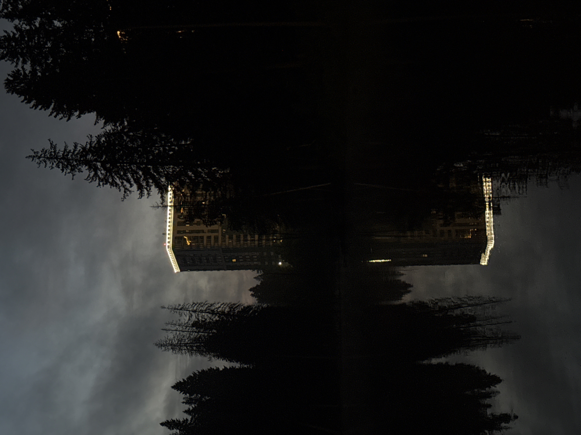 The image depicts a tall building with illuminated windows and a brightly lit top edge, situated behind a dark silhouette of pine trees. The building's reflection is visible in the dark, calm water in the foreground, which also reflects the trees and the sky. The sky is overcast with dark clouds, and the overall scene suggests either twilight or a dark, cloudy day. There are no people or animals visible in the image. This scene is likely located in Izhevsk, Russia, given the context of the query.