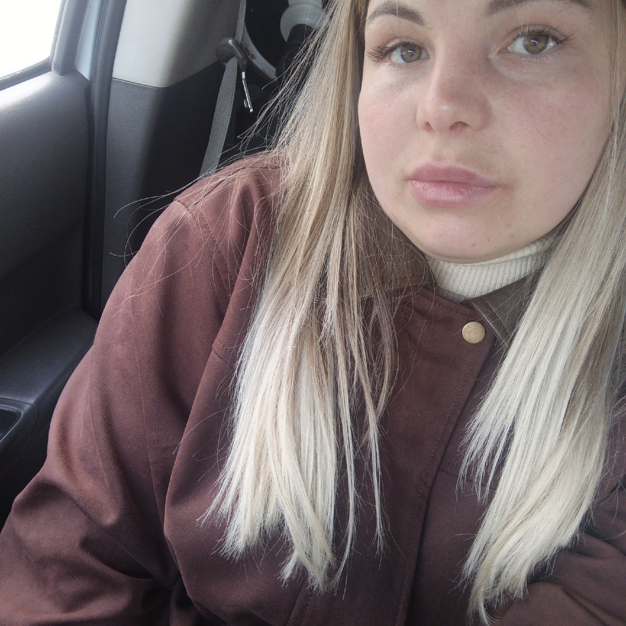 A woman is seated in a car. She has blonde hair and is wearing a brown jacket over a light-colored turtleneck sweater. A seatbelt is visible in the background. The car's interior is visible, including part of the door panel.