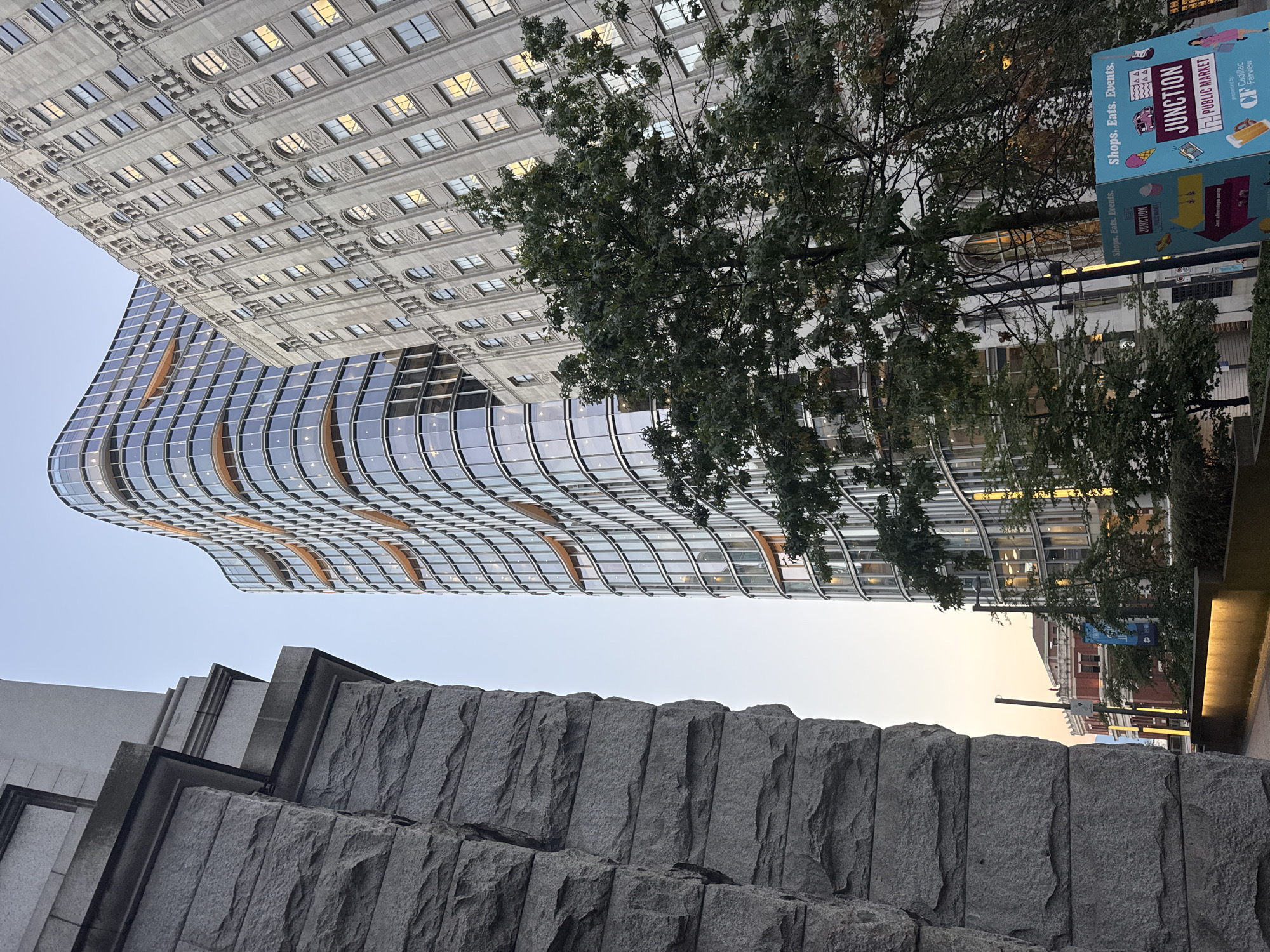 The image is a street-level view of a city intersection in Vancouver, Canada, featuring a mix of architectural styles and greenery. In the foreground to the left, a large, rough-hewn stone structure dominates the frame, with distinct horizontal layers of stone blocks. To the right of this stone structure, a modern, curved glass skyscraper with a distinctive wavy facade rises into the sky. The skyscraper's windows reflect the ambient light, and illuminated bands of orange light are visible between floors.
Behind the modern skyscraper, an older, ornate building with a grey stone facade and numerous arched windows is visible. Its windows also show reflections of interior lighting, suggesting the time of day is either early morning or late evening.
In the middle ground, a mature tree with dense green foliage partially obscures the older building and the modern skyscraper. Further back, another building with a red brick facade and ornate white trim can be seen, illuminated by yellow lights.
In the lower right corner, a teal-colored sign with graphics and text is prominently displayed. The text on the sign reads "Shops. Eats. Events. JUNCTION PUBLIC MARKET" and mentions "presented by Cadillac Fairview". This sign indicates the presence of a public market and points to directions with arrows.
The sky is a pale blue, with a subtle hint of warmth, suggesting a sunrise or sunset. The overall scene depicts an urban environment where new and old architecture coexist, with natural elements like trees integrated into the cityscape. The image is taken from a low angle, emphasizing the scale of the buildings against the sky.