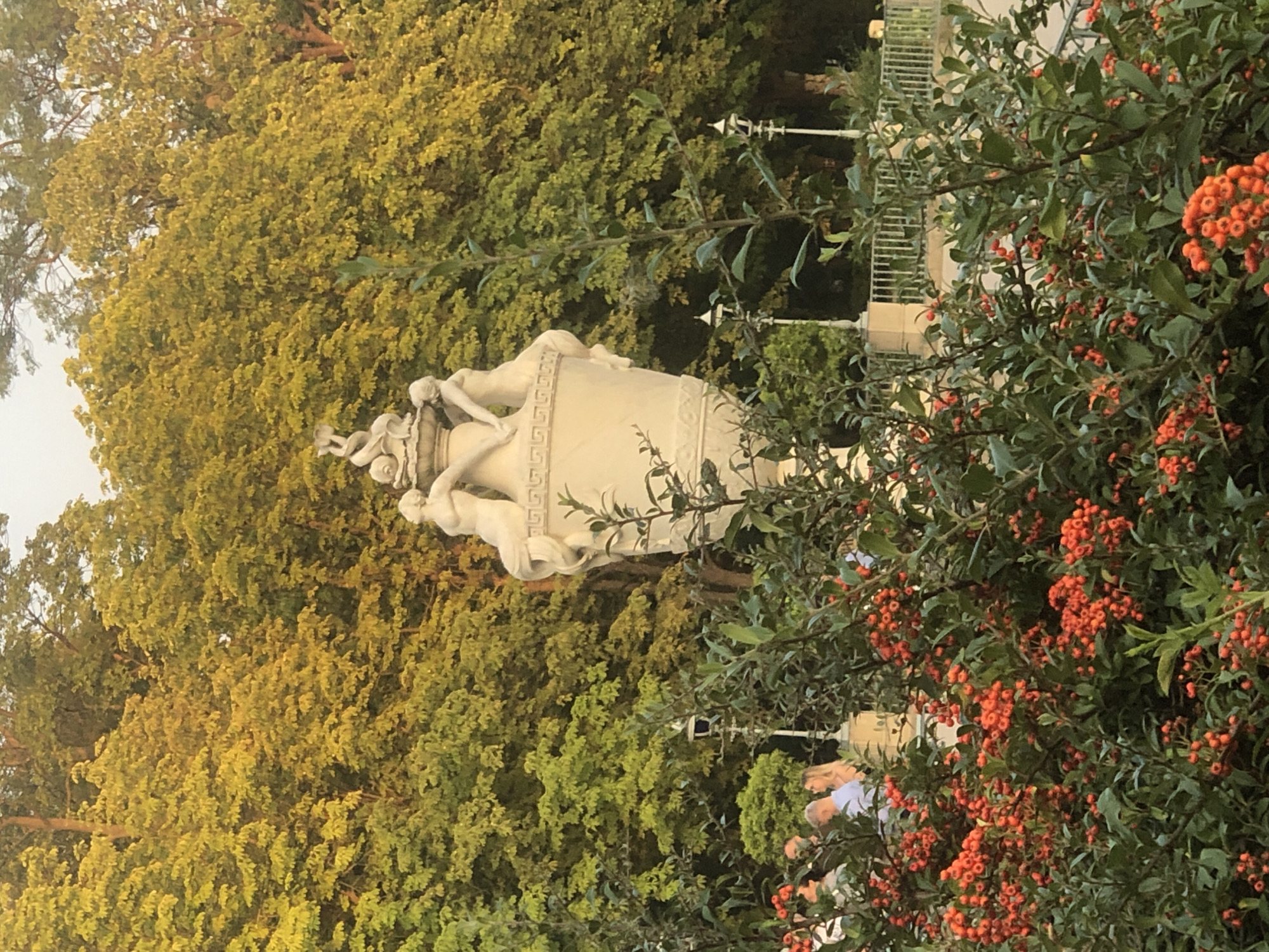 The image depicts a large decorative urn topped with sculpted figures, situated outdoors amidst lush greenery and trees. In the foreground, a dense bush laden with small, bright orange berries partially obstructs the view. Behind the urn, a formal garden setting is visible with lampposts, a railing, and several people in casual attire walking and conversing. The overall scene suggests a park or garden environment, possibly during daylight hours, with the warm lighting indicating either morning or late afternoon. The urn itself is a prominent, classical-style sculpture, possibly made of stone or plaster, featuring intertwined human figures. The trees in the background display a mix of green and yellow foliage, hinting at an autumn season or a particular type of tree. The presence of people suggests a public space. The location is identified as Łazienki Park in Warsaw, Poland.