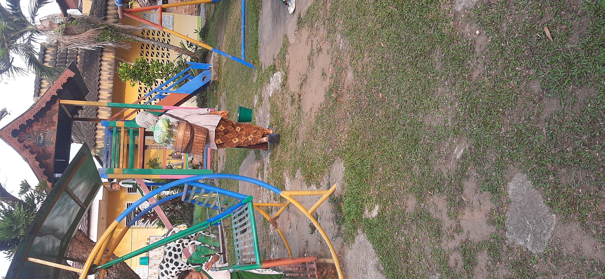 An outdoor scene at what appears to be a playground or schoolyard shows several children and an adult. On the left, a young child in a green and white uniform stands near a spotted decorative cow structure and part of a colorful swing set. To the right of the child, an adult walks away from the camera, carrying a woven basket on their back filled with bottles, and holding a green bucket in their right hand. The adult is wearing a headscarf, a striped shirt, and a patterned brown skirt. The ground is a mixture of grass and dirt patches. In the background, a yellow building with a distinctive patterned roof is visible, along with a palm tree and other playground equipment. The overall impression is of a warm, daytime setting. While specific details placing this in Jambi, Indonesia, are not explicitly visible within the image itself, such a setting would be consistent with environments found in the region.