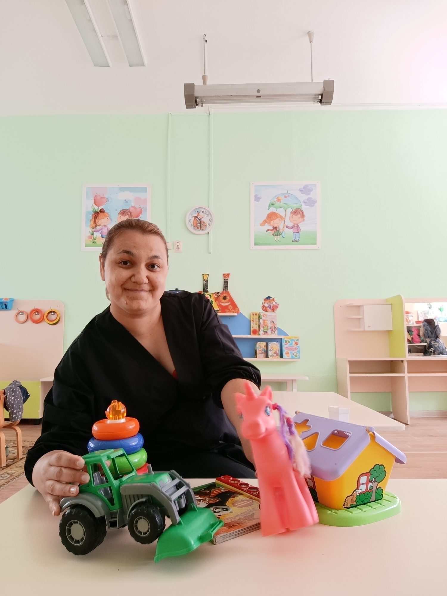 A woman with short, dark hair is seated at a light-colored table in a room with light green walls. She is wearing a black v-neck shirt. On the table in front of her are several children's toys, including a green toy tractor with stacking rings on top, a pink toy horse with purple hair, and a yellow and purple toy house. Behind the woman, on the wall, are two framed pictures and a small, round clock. A shelf unit with toys and other items is visible in the background, along with other tables and chairs. Based on earlier contextual information, this photo was taken in the city of Chaikovsky, Russia.