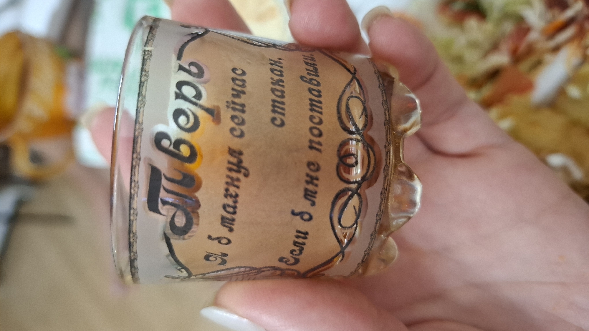 A person holds a small, ornate glass in their hand. The glass has a label written in Russian: "Пиверь. Я б махнул сейчас стакан, Если б мне поставили." This translates to "Pivér (a person's name). I would have a glass now if they put it in front of me." The glass also features a decorative pattern. The background is slightly out of focus, showing what appears to be food in a dish and other items on a table. The location cannot be determined from the image.