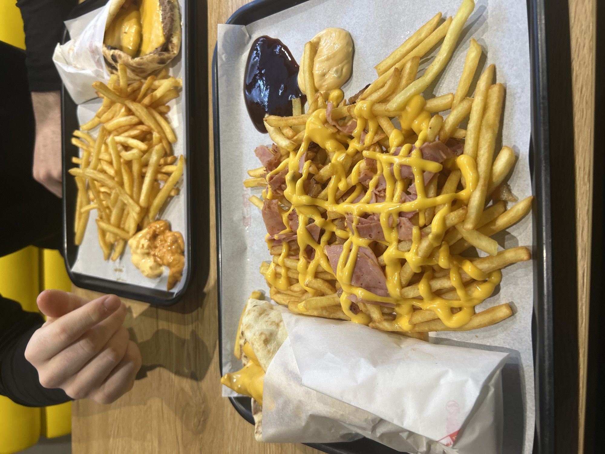 The photograph shows two black trays of food on a wooden table. The tray in the foreground holds a portion of fries covered with melted cheese and ham pieces. There is a sauce on the side. A wrap filled with what appears to be cheese and another ingredient, possibly meat, rests at the bottom of the tray. The other tray in the background features a burger wrapped in paper and a side of fries with a sauce. The hand of a person wearing a black shirt is visible in the lower-left corner of the image. The location appears to be Noisy-le-Grand, France.
