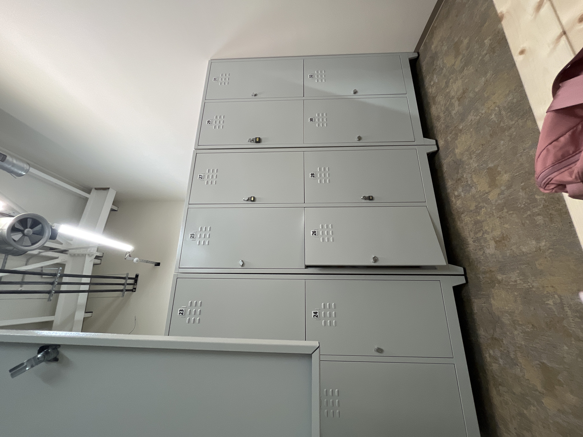 The image depicts a row of metal lockers in what appears to be a gym or changing room. The lockers are light gray and numbered sequentially from 23 to 32, with locker 26's door ajar. Some of the lockers have small padlocks on them. The floor is a distressed, wood-like linoleum. In the background, there are exposed pipes and industrial lighting fixtures on the ceiling. A pink duffel bag is partially visible in the foreground on a wooden surface. The scene is devoid of any individuals. The location is not identifiable as Plovdiv, Bulgaria, from the visual information provided.