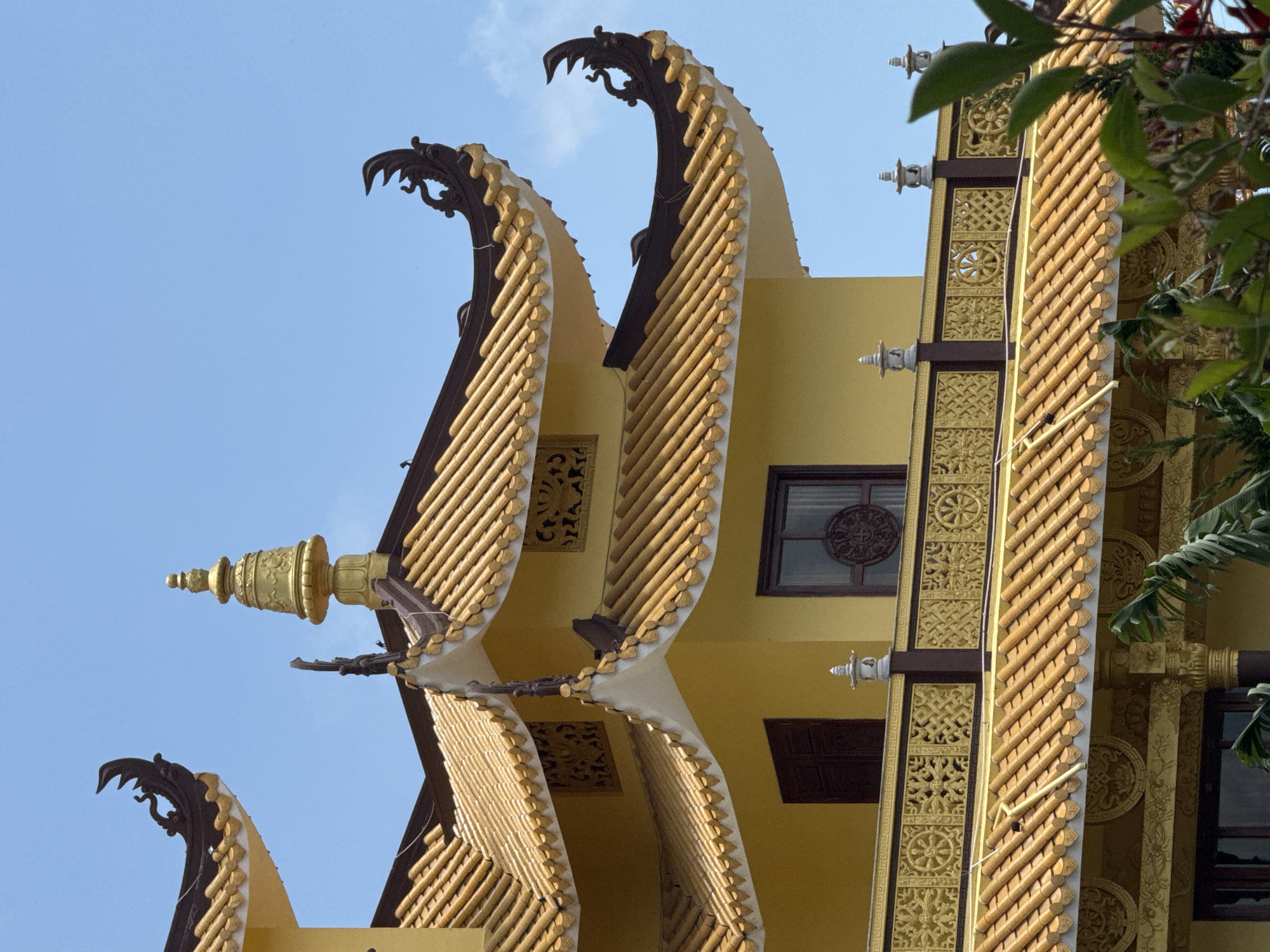 This is a close-up, eye-level shot of the distinctive yellow, multi-tiered, pagoda-style roofs of a Buddhist temple, likely the Thien Vuong Pagoda in Da Lat, Vietnam, under a clear blue sky. The architectural details of the temple are prominent: the curved, tiled roofs are adorned with intricate dark brown decorative elements, resembling stylized dragon heads or mythical creatures at the eaves. Below the upper roofs, a yellow facade is visible, featuring dark-framed windows, one with a circular, ornate design in the glass. A decorative railing with golden, relief patterns, including what appears to be Dharma wheels, runs along a balcony. Small, decorative pagoda-like finials sit atop posts along this railing. Green foliage is visible in the foreground and partially obscures the lower portion of the temple. The lighting suggests it is daytime, with no people or animals clearly visible.