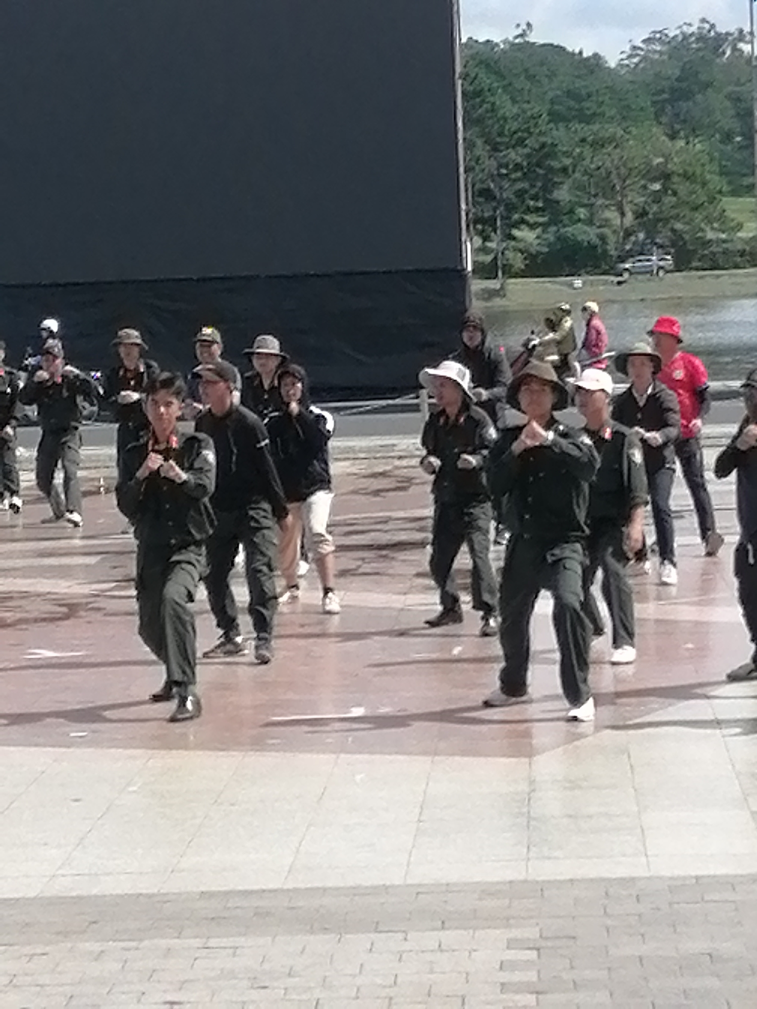 In Da Lat, Vietnam, a group of people, likely a martial arts or fitness class, are practicing outdoors on a paved area in front of a large, dark screen. The participants, predominantly male, are dressed in dark, uniform-like clothing, with some wearing hats. The scene captures a moment of synchronized movement, with individuals in various stances, suggesting a physical activity. The environment includes a backdrop of trees and a cloudy sky, indicating an outdoor urban setting. The lighting suggests it is daytime.