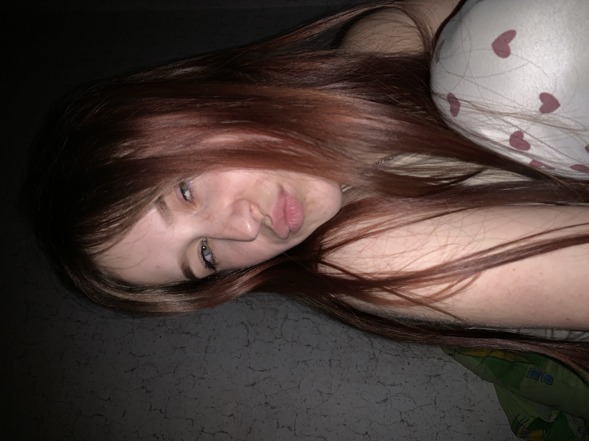 A woman with auburn hair is lying down, partially on a pillow with pink heart patterns, and partially on a surface that appears to be stone or concrete. The location is indeterminate within the city of Фоки, Russia. The woman's face is visible, and she is looking directly at the camera with a slight expression. Her arm is visible, resting on the surface, and a green and yellow object, possibly bedding, is visible at the bottom of the image. The lighting is subdued.