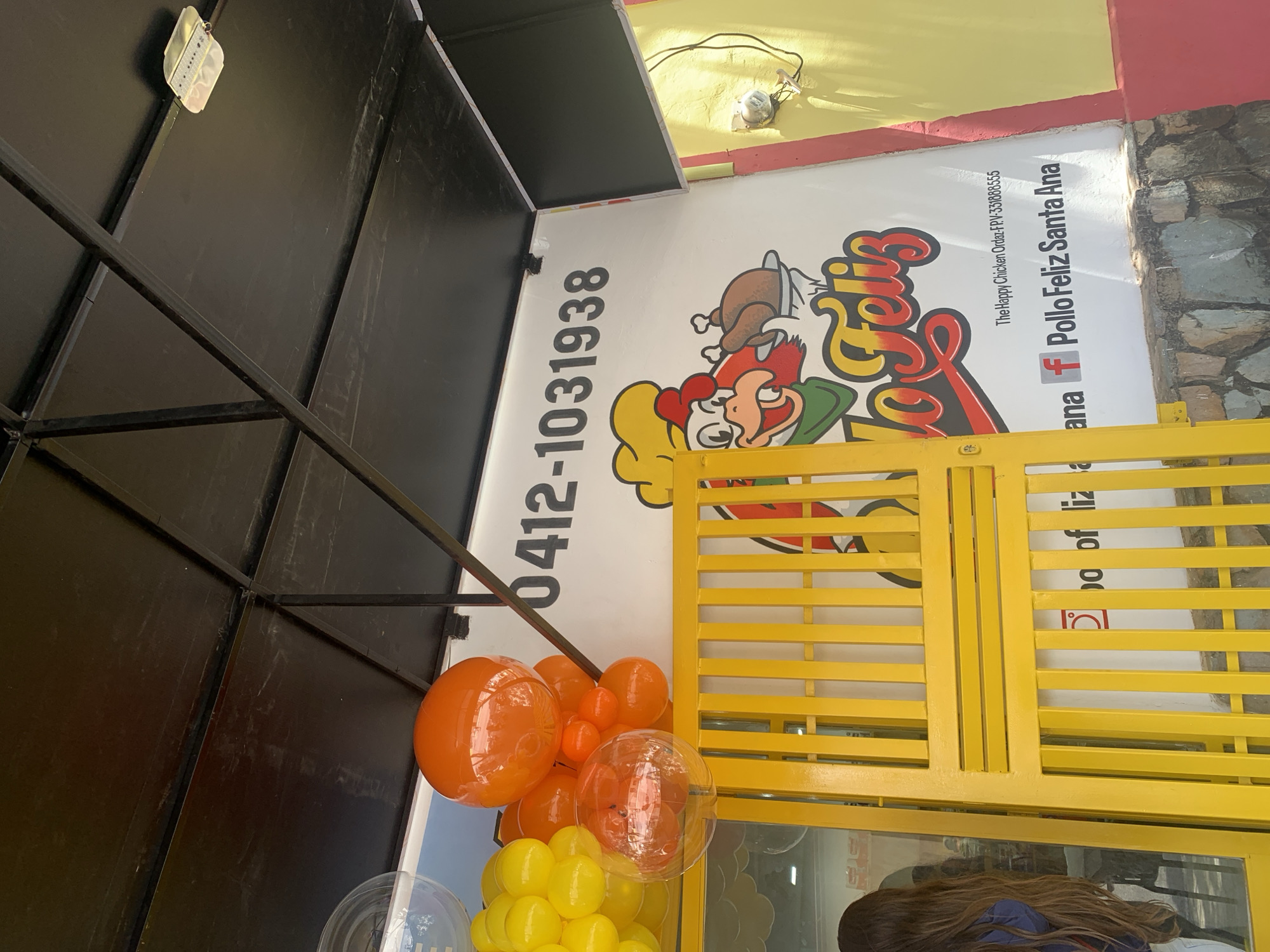The image depicts a partial view of a building with a sign for "Pollo Feliz Santa Ana," a chicken restaurant in Santa Ana, Venezuela. The phone number "0412-1031938" is prominently displayed on the sign, along with the logo of a cartoon chicken character. A string of orange and yellow balloons adorn the entrance. A yellow slatted barrier is visible, partially obscuring a person with long brown hair wearing a blue shirt. The background features a wall with a rough, stone-like texture. The framing of the picture gives an oblique view of the building's corner and architectural elements.