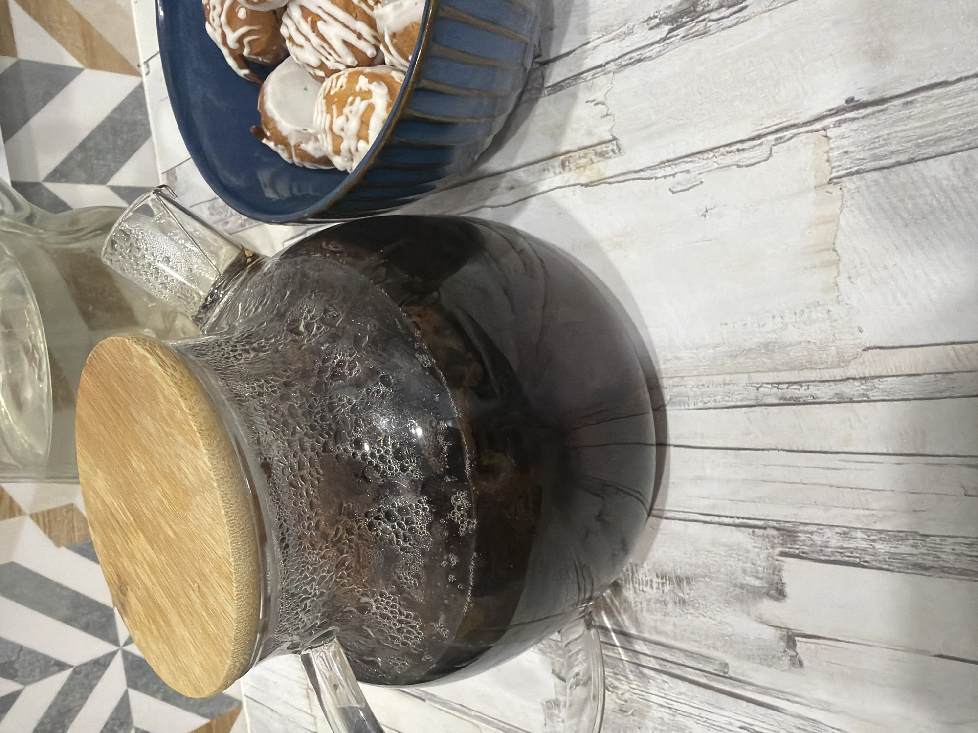 A close-up, high-angle shot shows a glass teapot with a wooden lid filled with dark liquid, likely tea or coffee, sitting on a light-colored, wood-patterned surface. Condensation is visible on the outside of the teapot. To the right, a blue bowl contains several round, fried pastries drizzled with white icing. In the background, a patterned tile backsplash is partially visible. The scene appears to be indoors, likely on a countertop or table. There are no people or animals present. The environment is clean and well-lit. There is no information available to place this image in Nizhnekamsk, Russia.