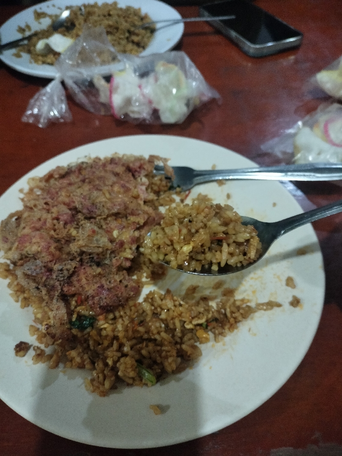 The image displays a close-up view of a casual meal setting, likely taking place indoors, with a focus on two plates of food on a reddish-brown table. The scene is set in Dramaga, Indonesia, based on the provided context.
In the foreground, a white ceramic plate holds a generous serving of what appears to be fried rice (nasi goreng). A significant portion of this rice is topped with a crumbly, reddish-brown ingredient, possibly shredded meat or corned beef. A metal fork is visible, resting on the plate near the topping. A metal spoon, held aloft over the plate, is filled with a spoonful of fried rice, suggesting that the meal is actively being eaten or is about to be consumed.
In the background, slightly out of focus, another white plate is visible, also containing fried rice, some of which has been eaten, and what appears to be a sliced hard-boiled egg. Cutlery is also present on this plate. Scattered on the table around the plates are several small, transparent plastic bags containing various colorful crackers (kerupuk), which are common accompaniments to Indonesian meals. A dark-colored smartphone is partially visible on the table in the upper right section of the image.
The lighting is dim, indicating either an evening meal or an indoor setting with soft artificial illumination. There are no people visible in the frame, and no visible text can be discerned on any objects. The overall impression is one of a simple, local dining experience.