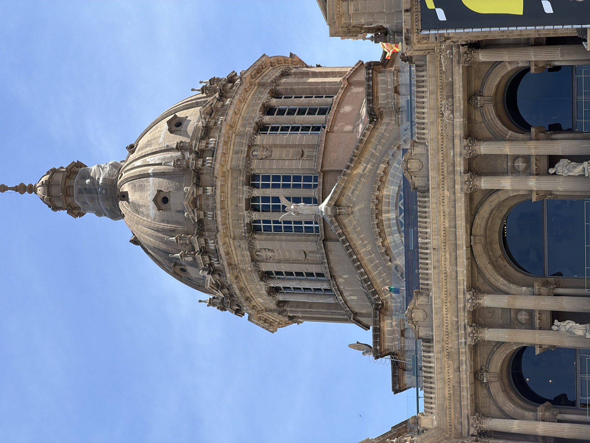 The image shows a close-up, low-angle view of the National Palace of Montjuïc in Barcelona, Spain. The palace, an imposing neoclassical building, is dominated by a large dome with intricate architectural details and ornate windows. Several statues are visible on the facade. In the foreground, a modern glass barrier and a black banner with a yellow design are present, suggesting a visitor area or an event. Two people are visible on a balcony area, one wearing a green shirt and blue shorts, and another in the distance. The sky is a clear, bright blue. The National Palace is located on Montjuïc hill in Barcelona, not in L'Hospitalet de Llobregat.