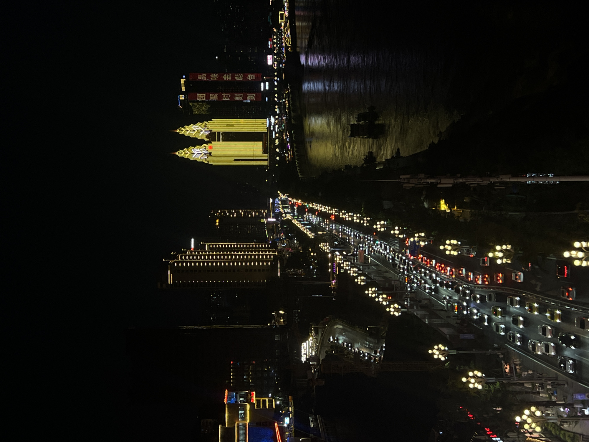 The image depicts a nighttime cityscape in Nan'an District, Chongqing, China, overlooking a busy highway bridge spanning a wide river. The highway is heavily congested with traffic, characterized by rows of illuminated vehicles, their headlights and taillights creating streaks of light against the dark asphalt. Ornate streetlights, each with multiple glowing bulbs, line the sides of the bridge, illuminating the roadway and the adjacent pedestrian walkway.
On the left side of the bridge, several buildings are visible. One prominent structure features a yellow facade with continuous vertical lines of lights. Further left, a larger building complex exhibits colorful illuminated signage and architectural lighting. On the right side of the river, the skyline is dominated by several tall buildings. Two particularly striking buildings are illuminated with bright yellow lights, resembling twin towers with pointed, illuminated spires. Adjacent to these, a red banner with white Chinese characters is displayed vertically on a building.
The river itself reflects the city lights, creating shimmering patterns on its surface. A few boats or barges are visible on the water, their lights also contributing to the illumination. The overall scene is one of urban vibrancy and nighttime activity, with a strong emphasis on the illuminated architecture and the flow of traffic.