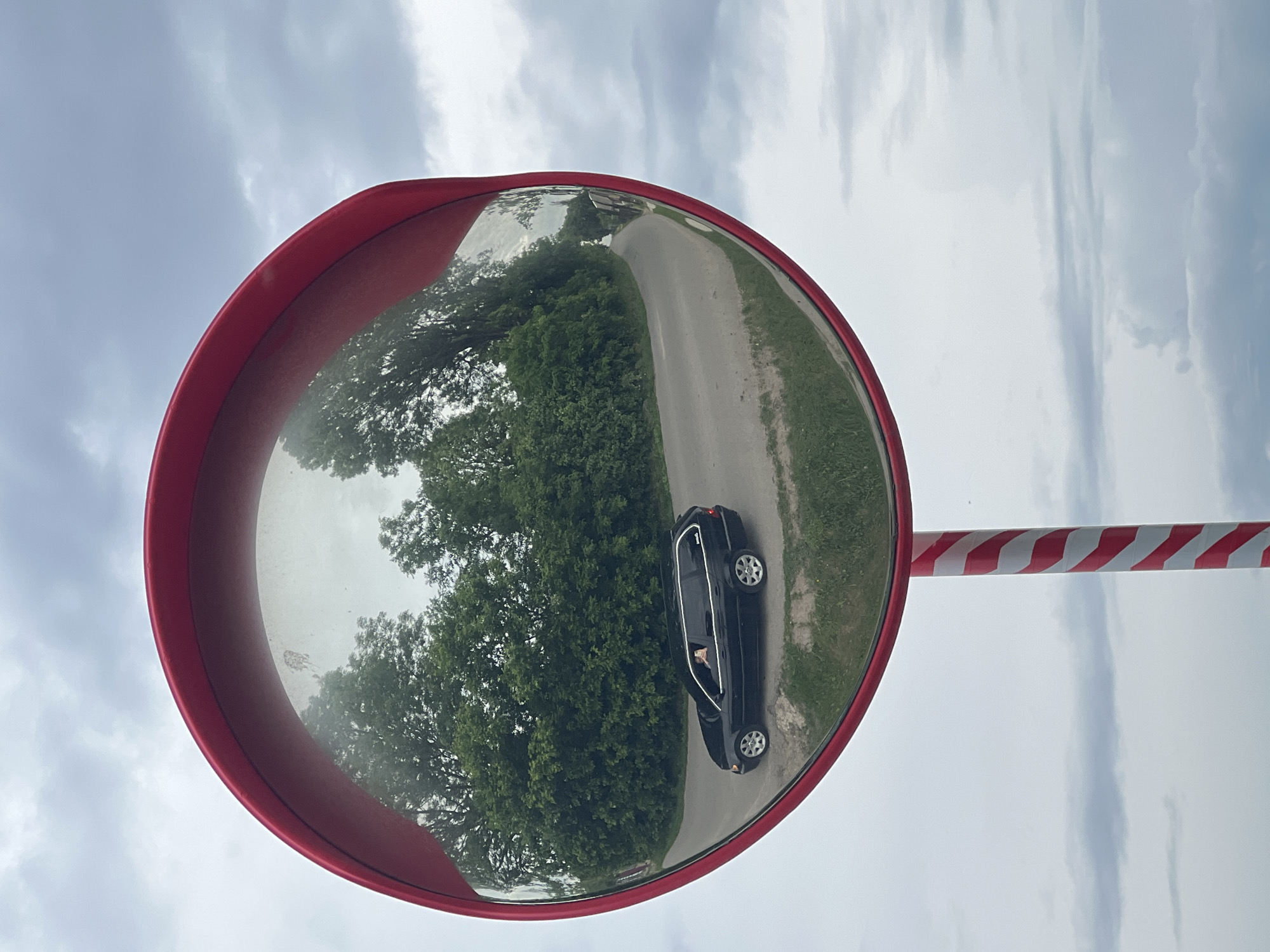 A convex traffic mirror with a circular red frame is positioned on a red and white striped pole in Kozivka, Ukraine, under an overcast sky. The mirror reflects a black station wagon, possibly a BMW E46, navigating a curved, paved road. An individual's arm extends from the front passenger window of the vehicle, holding an object. The road is bordered by dense green trees and grass. The reflection also captures the light grey, cloudy sky. A faint, irregular smudge is visible on the upper left portion of the mirror's surface. The background outside the mirror consists solely of a cloudy sky with varying tones of grey.