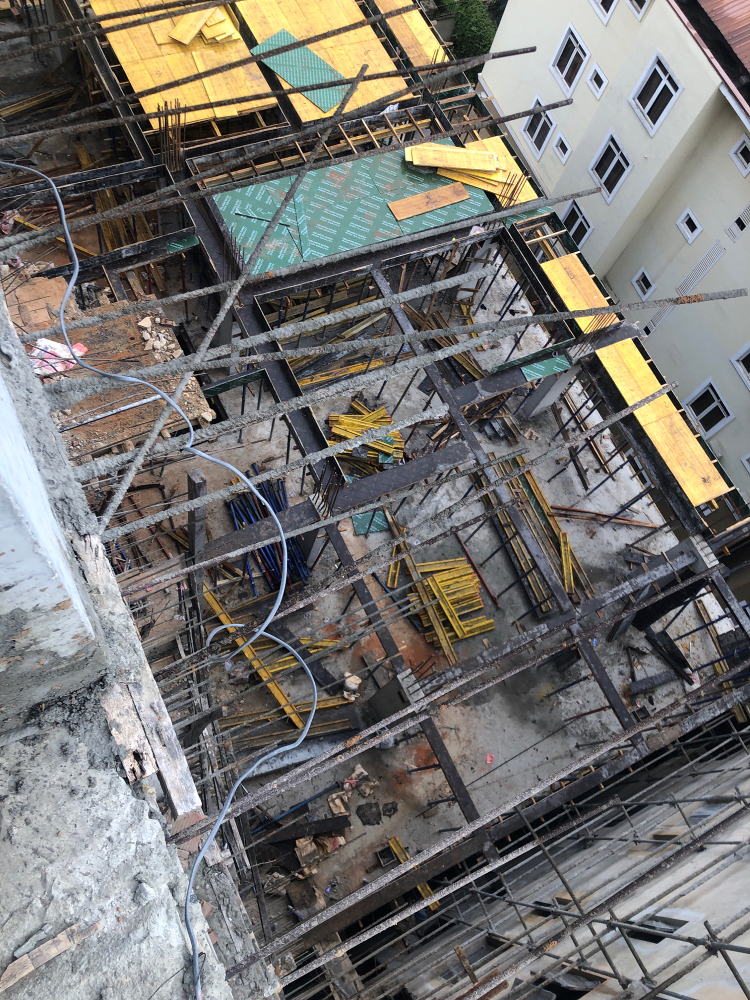 An overhead, angled view shows a construction site for a multi-story building in Ikeja, Nigeria. The partially completed structure reveals a network of concrete, rebar, and wooden scaffolding. Yellow and green construction materials are visible on various levels, along with stacks of blue pipes. The scene is characterized by unfinished construction, with exposed rebar and rough concrete surfaces. A light-colored, multi-story building with numerous windows stands adjacent to the construction area, suggesting an urban setting. No people are depicted in the image. The lighting indicates it is daytime.