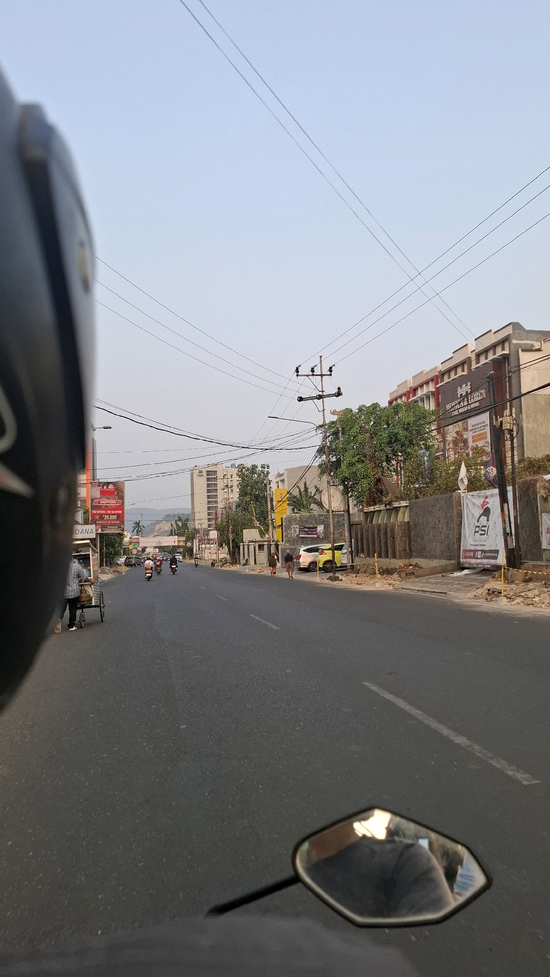 A first-person perspective shot from a motorcycle on a wide, paved road in Bandar Lampung, Indonesia. The road is lined with buildings and trees. On the right side of the road, there's a prominent building with signs for "ANUGRAH & LOREN" and "PSI". A yellow car is parked in front of a building. On the left side of the road, there are several motorcycles, a food cart with a vendor, and a billboard. Power lines crisscross the sky. The sky is clear and light blue, indicating it's daytime, likely late afternoon or early morning. The overall scene depicts a typical street view in an urban environment.