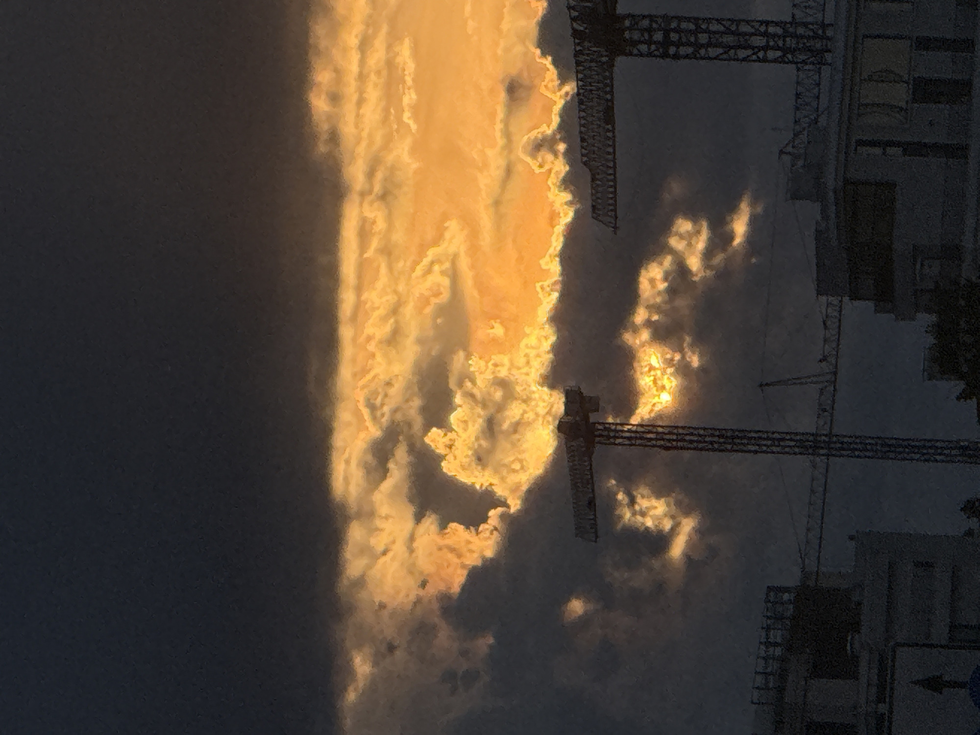 The image captures a dramatic sky with brightly lit clouds, appearing to be illuminated by the setting sun. The clouds are a vibrant orange and yellow, contrasting with the darker, overcast sky. Silhouetted against this striking sky are the structures of construction cranes and buildings. One crane's latticework is clearly visible, extending upwards. The scene suggests an urban setting undergoing development. The time of day is clearly evening, given the intense coloration of the clouds, indicative of sunset. The weather appears to be partly cloudy, with pockets of intense light breaking through. No people are visible in the frame. The overall mood is one of awe at the natural spectacle of the sky, juxtaposed with the industrial elements of construction. There is a small blue sign with a white arrow pointing left, a subtle location cue.