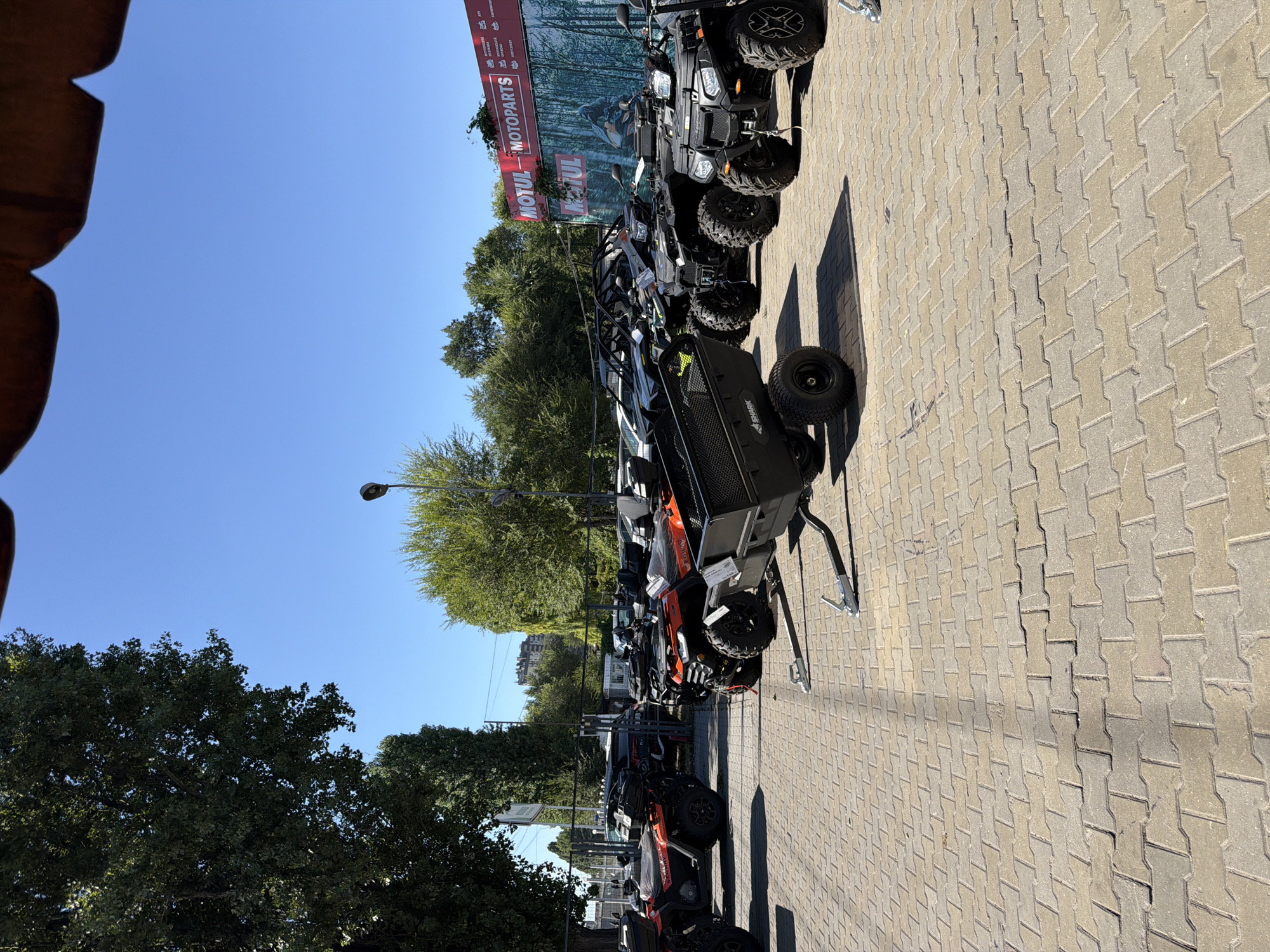 The image depicts an outdoor exhibition of ATVs and a small trailer parked on a paved lot in Chișinău, Moldova. The ATVs are predominantly orange and black, with several models from the Segway brand visible. A black trailer with "SHARK" branding is positioned in the foreground, attached to an unseen vehicle. In the background, a large banner for "MOTUL - MOTOPARTS" is displayed, suggesting the location is a dealership or a related automotive service. Trees line the perimeter of the lot, and the sky is clear and blue. There are no people visible in the image.