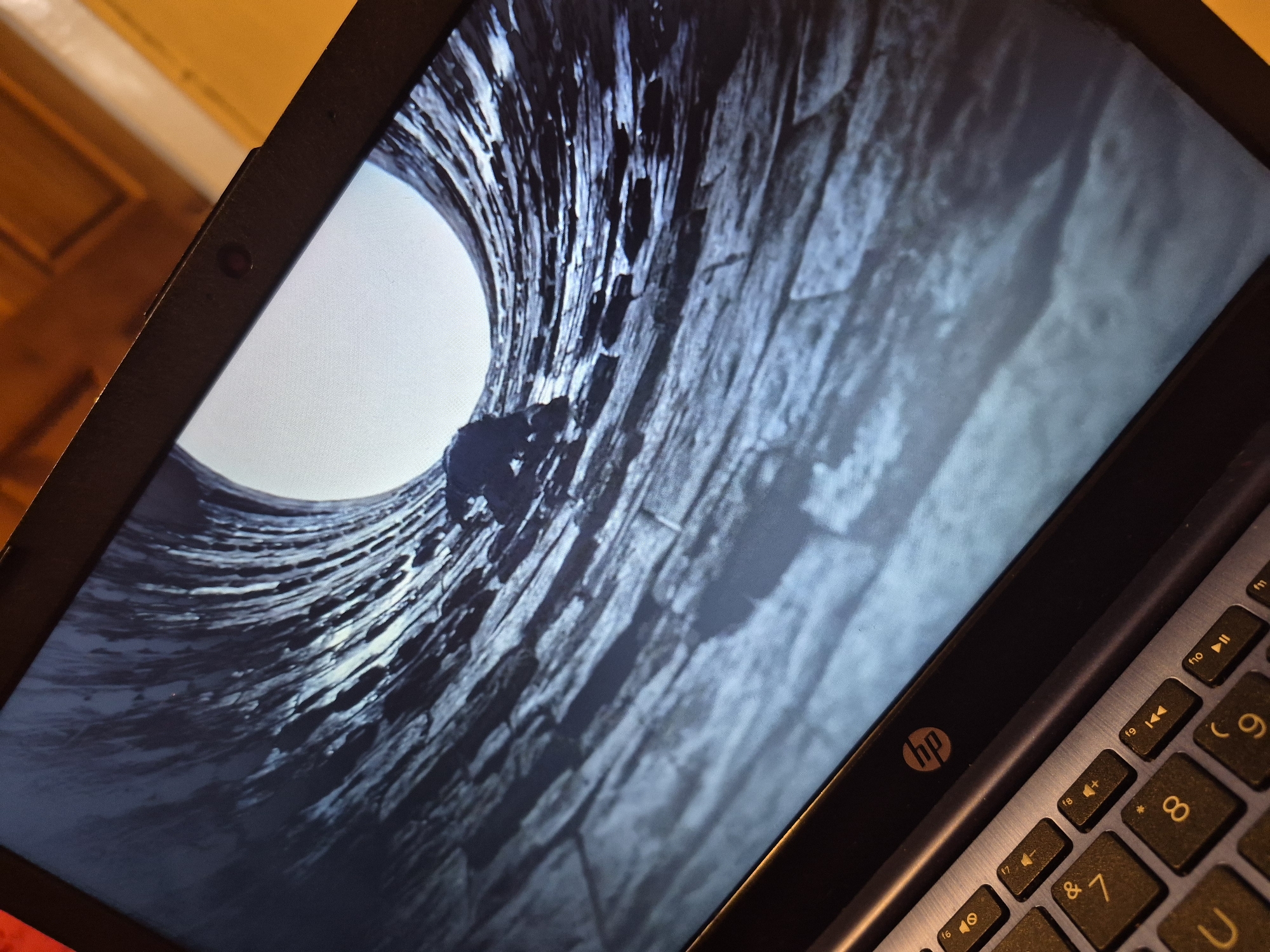 A laptop is open, displaying a dark, textured image of a person climbing out of a cylindrical well. The well's interior appears to be constructed of roughly hewn stones, with the opening at the top letting in bright light. The person is silhouetted against the light, appearing to be pulling themselves upwards. The laptop itself is dark, with a visible HP logo above the keyboard. The keyboard has yellow lettering on black keys. The scene is dimly lit, with warm orange light reflecting from the background onto the laptop. The location is not identifiable within the city of Sandwell, United Kingdom.