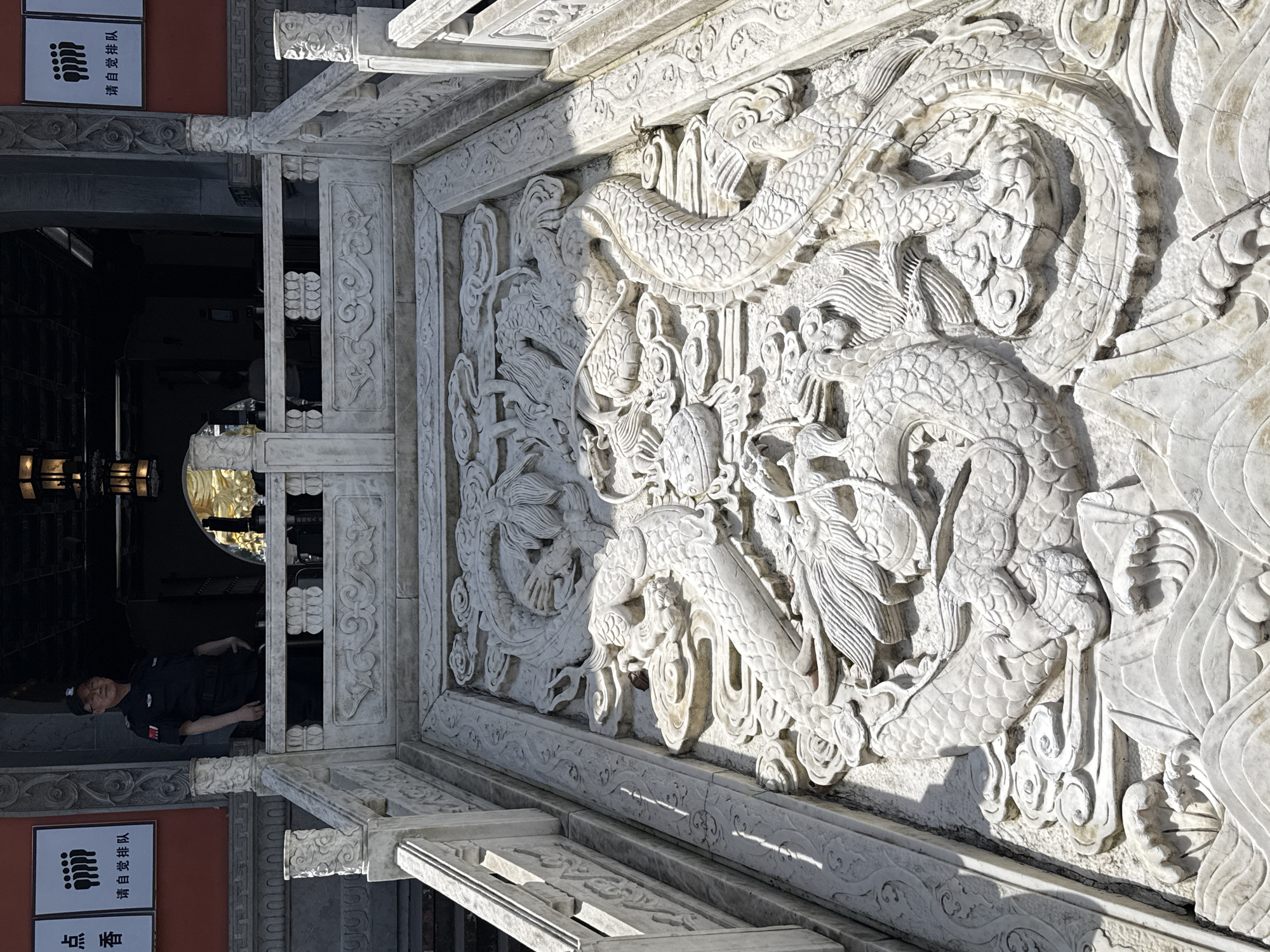 The image shows a detailed, intricately carved white stone wall with a relief of intertwined dragons, located in Jiulongpo District, China. A portion of a building’s entrance is visible to the left, including a security guard in a dark uniform standing inside the entrance. A sign with Chinese characters and an image of several silhouettes of hands is posted near the door. Light is cast across the dragon relief, creating distinct shadows that accentuate the carvings.