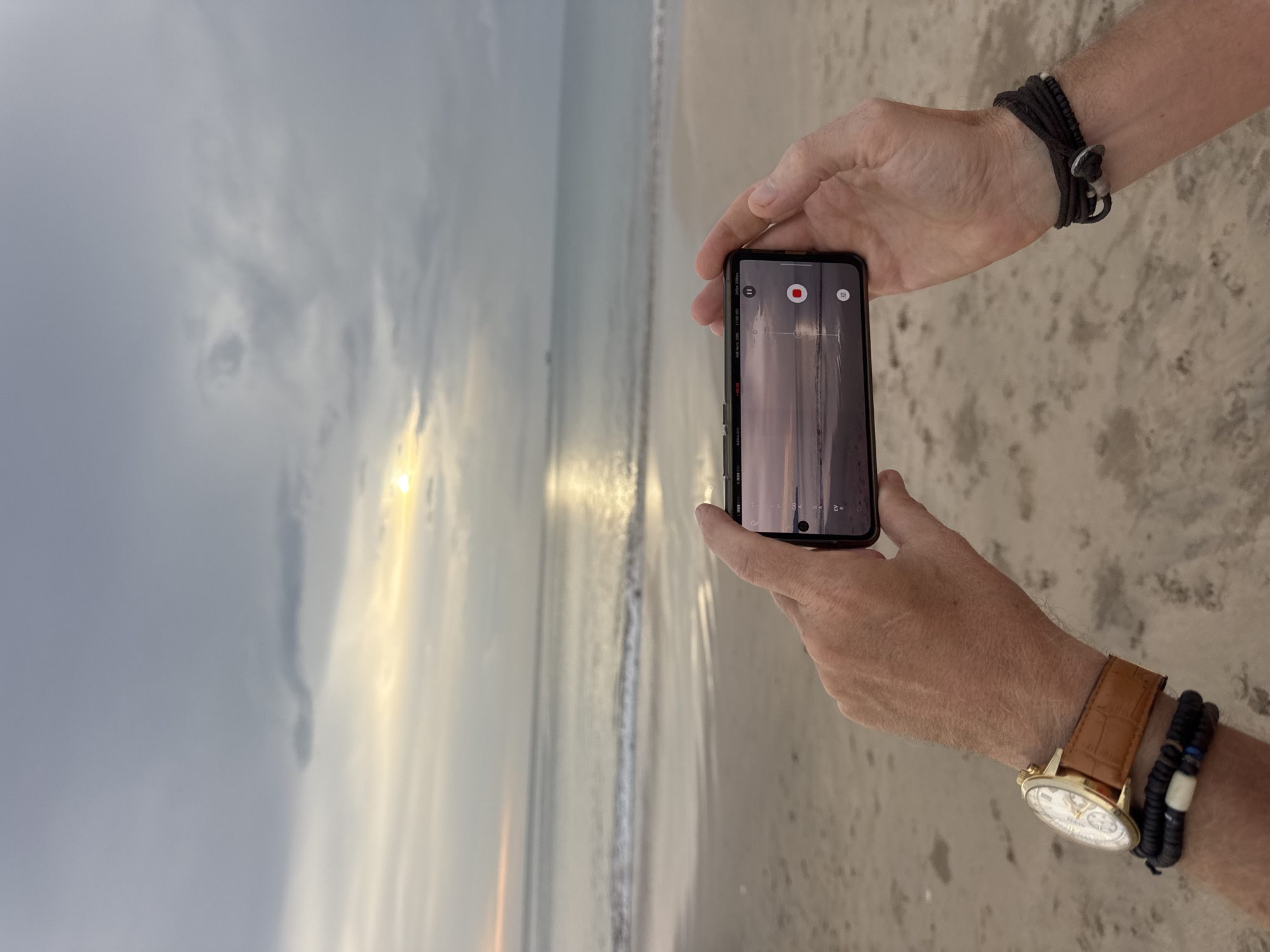 An individual's hands hold a black smartphone horizontally, capturing a picturesque beach scene. The screen of the phone displays a camera interface, including a red "REC" button and a timer, suggesting video recording is in progress or paused. The phone's camera is pointed towards the ocean and sky, mirroring the broader vista.
The setting is a sandy beach in Kuta Selatan, Indonesia, with the ocean stretching out under a cloudy sky. The sun, low on the horizon, casts a bright, golden reflection across the water, indicating either sunrise or sunset. The clouds are a mix of light and dark grey, with the sun's light breaking through in places, creating a dramatic atmosphere.
The person's forearms and hands are visible. On the left wrist (viewer's right) is a dark, multi-strand bracelet with beads and a silver-toned clasp. On the right wrist (viewer's left), a gold-colored wristwatch with a light-faced dial and a brown leather strap is worn alongside a black and white beaded bracelet. The focus is on the act of digitally preserving the serene coastal view.