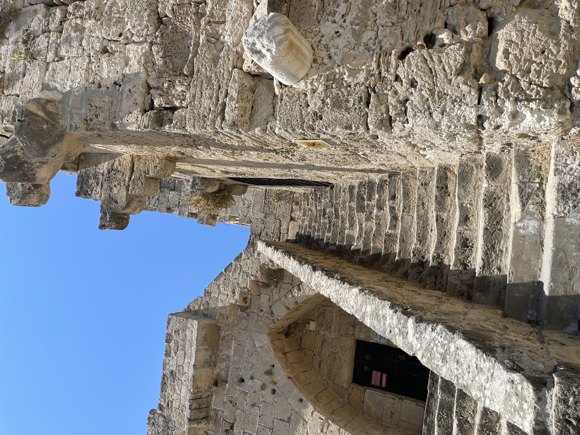 This is a low-angle, close-up, outdoor shot of a stone staircase leading up to an arched doorway within an ancient stone structure. The stonework is rough and weathered, with various sizes of irregular blocks. A thick, textured stone railing runs along the right side of the stairs. The doorway appears to be made of dark wood and has some signs attached. The sky above is a clear, bright blue. The overall impression is one of historical architecture, possibly a castle or fortification, set against a sunny sky. It is likely located in Kyrenia, Northern Cyprus.