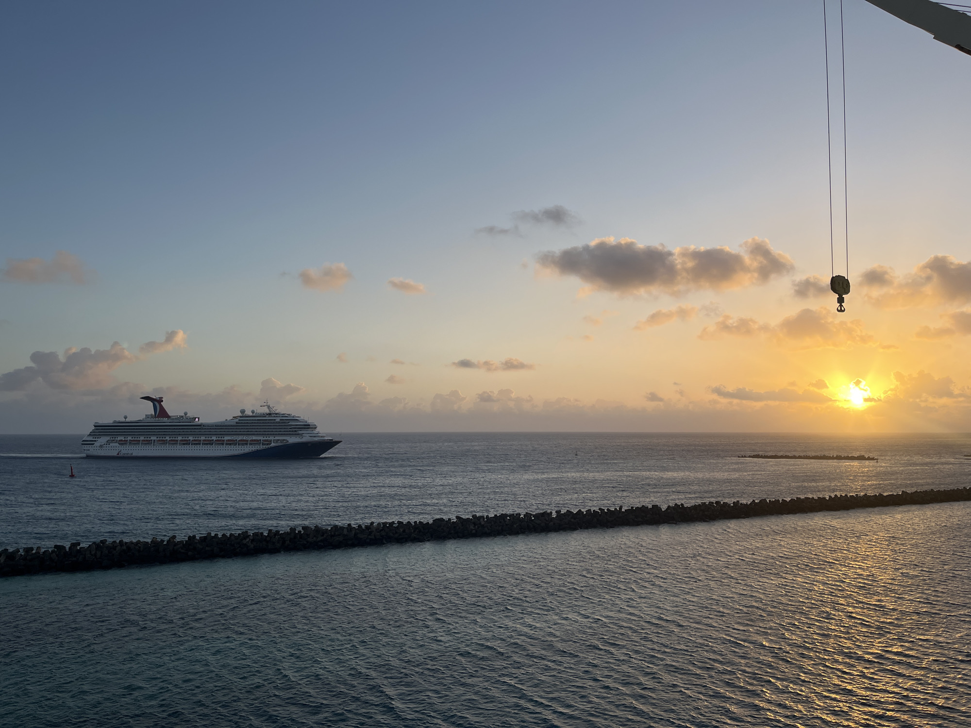 The image captures a cruise ship, marked with a red and white logo, cruising on the ocean near a breakwater. The sun is setting or rising, casting an orange glow across the clouds and the water. A crane's hook hangs from the upper right, suggesting a port or construction area nearby. The location is likely a coastal area, possibly near the city of Unknown, Unknown, but specific details are not available.