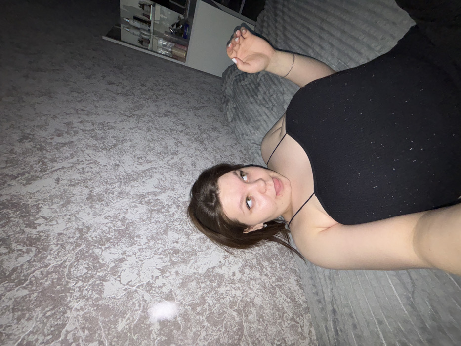 The image displays a single female individual within an indoor residential setting; the specific location in Perm, Russia, is not ascertainable from the visual evidence. The subject, a young adult female with medium-length brown hair, is clad in a black spaghetti-strap top. She is depicted in a reclining position on a grey, textured fabric surface, likely a bed or sofa. Her left arm is extended towards the camera, suggesting a selfie. Her head is tilted to her left, with her eyes directed upwards and to the right of the frame. Her mouth is slightly open, and her tongue is subtly protruded, conveying a playful or whimsical expression. Her right arm is bent at the elbow, with the hand partially visible and fingers slightly splayed, resting on the surface. The background features a wall covered in a greyish-white textured wallpaper. To the far right, a white shelving unit, populated with various bottles and containers, is partially visible.