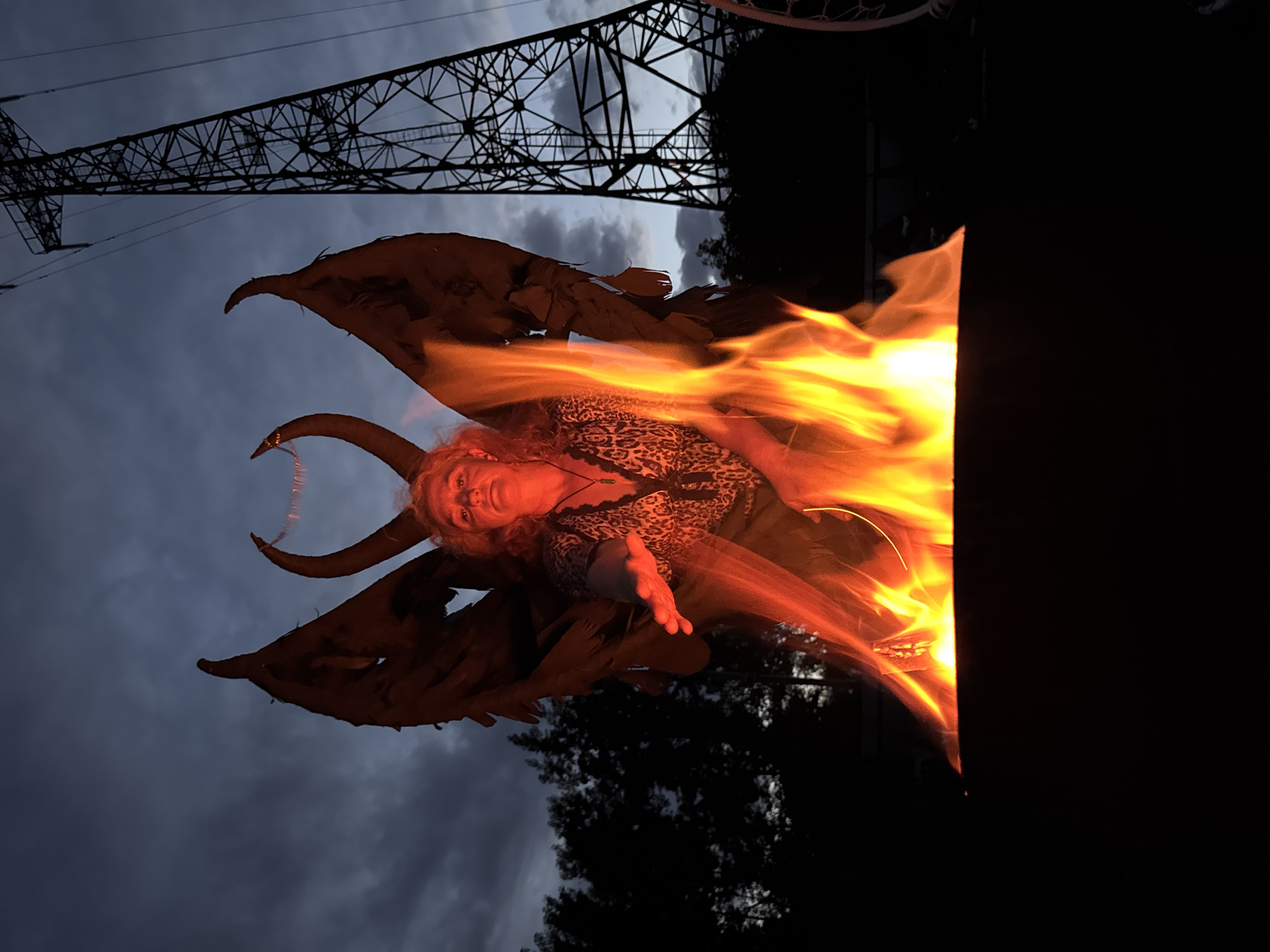 A woman with curly blonde hair, wearing a leopard print top and a dark skirt, is positioned in front of a large fire. She has a costume on with large, dark, feathered wings and prominent, curved horns. One of her hands is extended forward. The fire is in a dark cauldron or pit, with flames reaching up around the woman. Behind her, a tall, dark electrical pylon dominates the background. The sky is a dusky blue with dark clouds. The scene appears to be outdoors, possibly in a rural or semi-rural area, with silhouetted trees visible to the left and a power line structure to the right. The overall impression is one of a dramatic, possibly staged, scene with a theme of supernatural or mythical figures. The location within the city of Chaikovsky, Russia is not discernible from the visual information provided in the image.