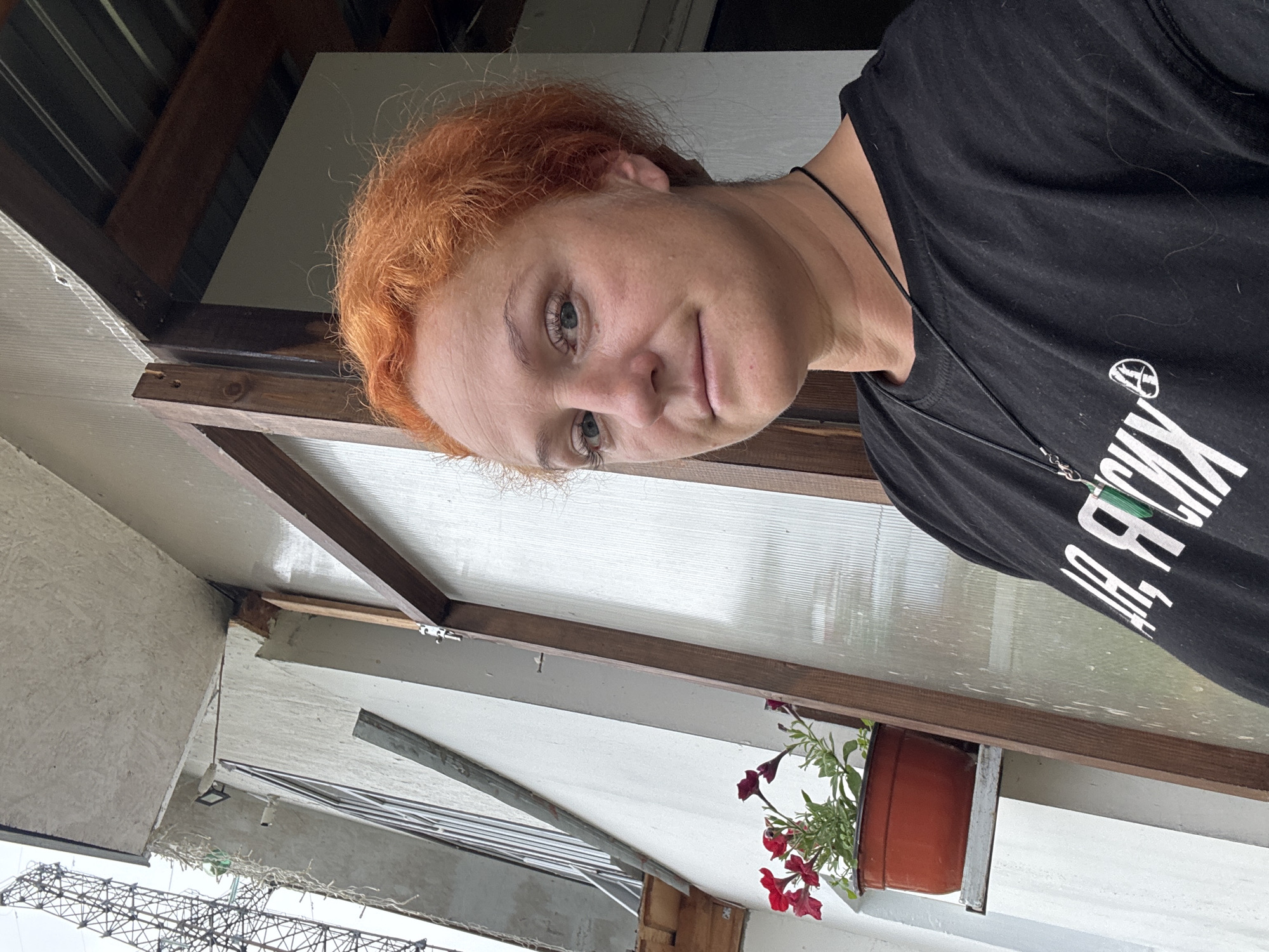 A medium shot selfie shows a fair-skinned Caucasian woman with short, bright orange hair and light blue eyes. She is wearing a black t-shirt with white lettering and a black cord necklace with a pendant. Her head is turned slightly to the right, and she is looking directly at the camera with a neutral expression.
The background suggests an outdoor setting, likely a porch or balcony area. To the left, a white wall with a window is visible, and a red flower pot with blooming red flowers sits on a shelf attached to the wall. Above and to the left of the woman, a metal structure resembling a radio tower or utility pole is partially visible against an overcast sky. To the right of the woman, a wooden frame surrounds a translucent, corrugated panel, part of a structure like a greenhouse or covered walkway. The overall impression is of an everyday scene in a residential area.
While the exact location cannot be determined from the visual information alone, the context of Chaikovsky, Russia, suggests this could be a typical residential exterior in a town known for its industrial development and surrounding natural landscape. The presence of the flower pot and the rustic wooden structure points to a personal, perhaps rural or semi-rural, setting.