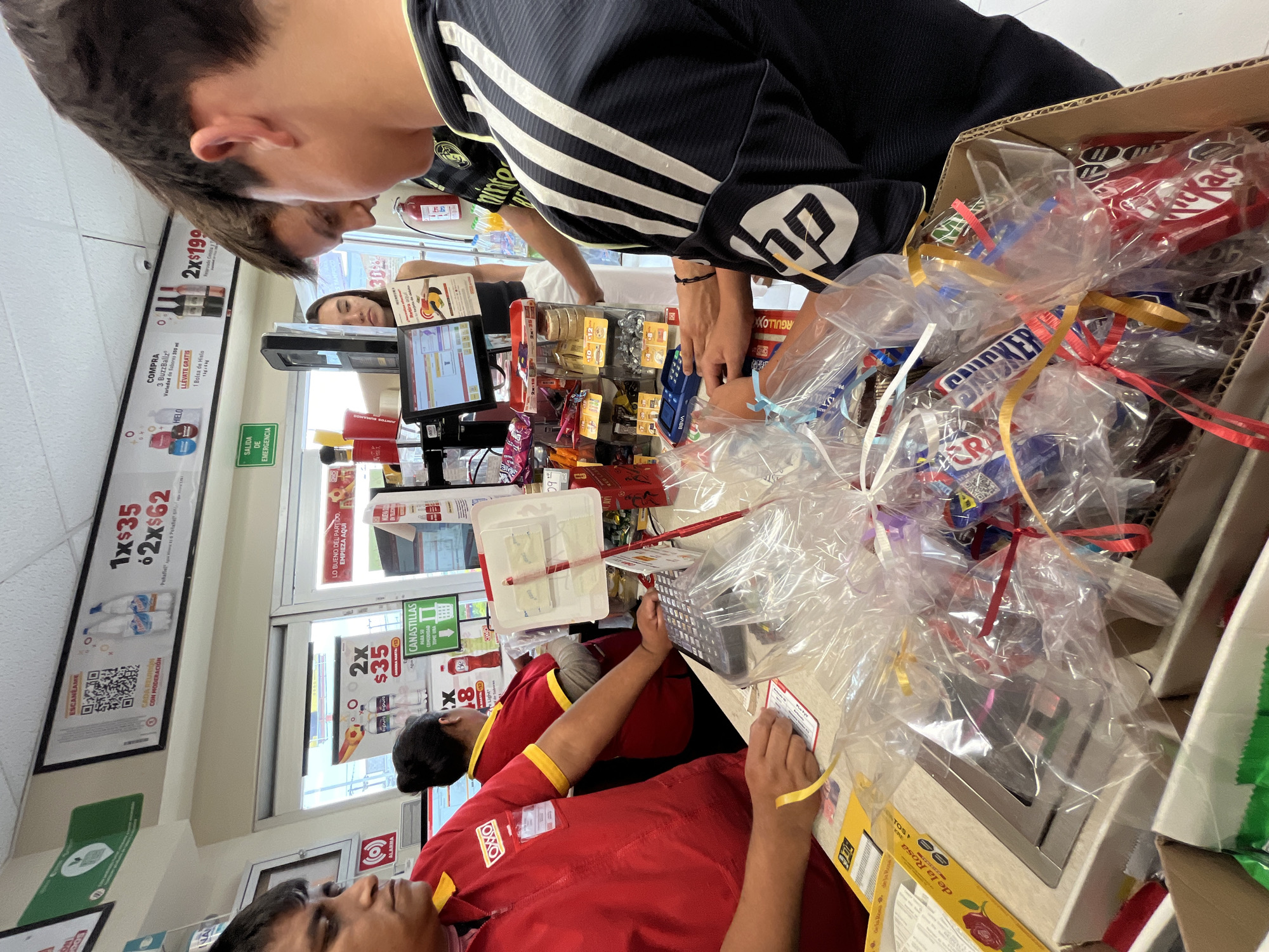 This is an interior shot of an OXXO store checkout in San Bernardino Tlaxcalancingo, Mexico. Two employees are present, both wearing red OXXO uniforms with yellow trim. One is operating the cash register, while the other is facing forward, seemingly assisting a customer. A young man in a black and white striped athletic jacket is visible, presumably a customer. In front of him is a cardboard box filled with individually wrapped candy, adorned with ribbons. The checkout area is cluttered with various items, including a point-of-sale system with a screen displaying transaction details. Walls are covered with promotional advertisements for products like water and alcohol. A security camera is mounted on the ceiling near signage.