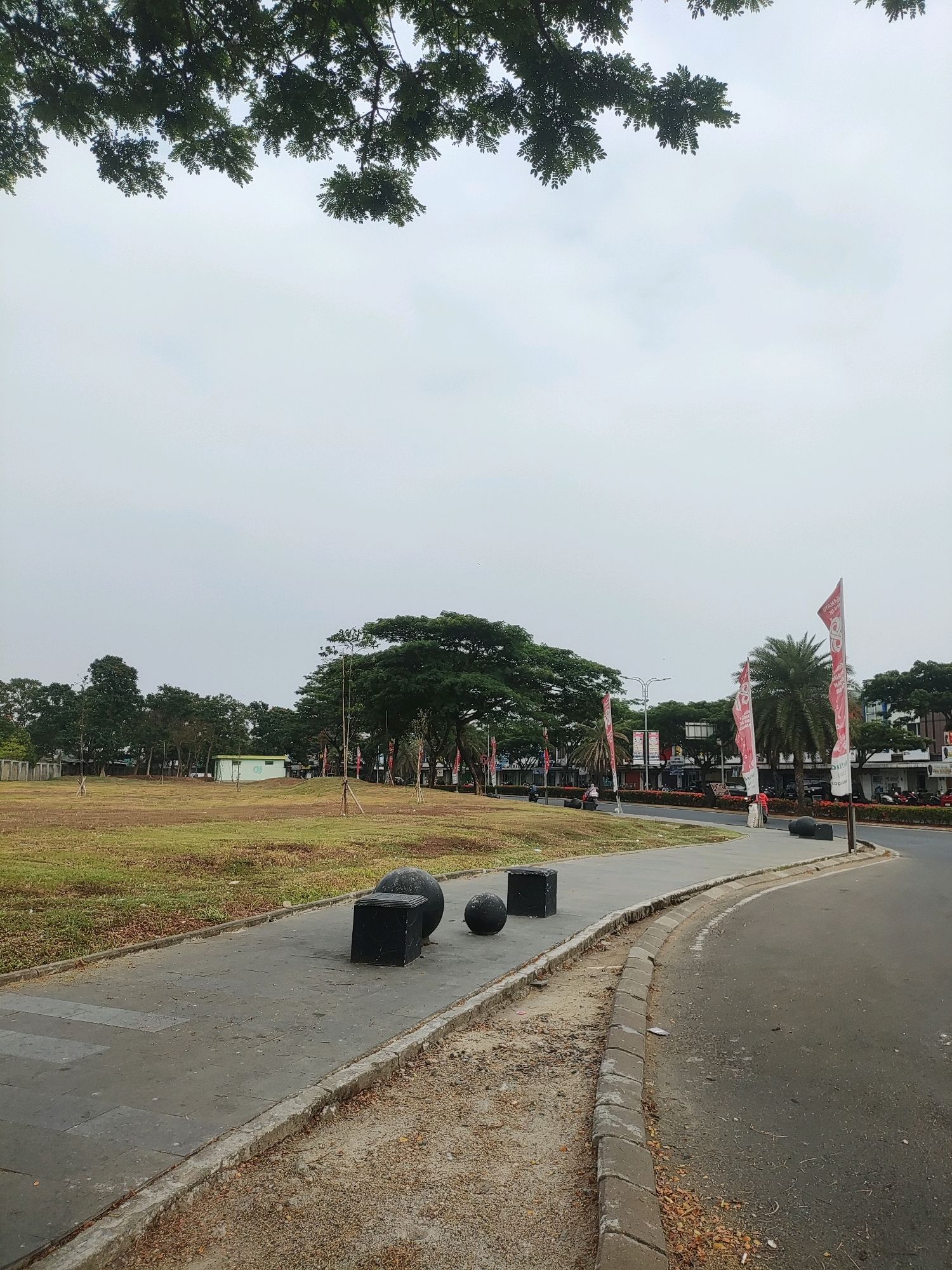 The image depicts an outdoor scene on an overcast day, likely in Telagaasih, Indonesia. In the foreground, a curved asphalt road runs alongside a paved walkway. On the walkway, three dark, geometric sculptures are placed: one cube and two spheres. Beyond the walkway, a large, open grassy field stretches out, dotted with young trees staked to the ground. In the middle ground, a small, light-colored building with green accents stands in the field. Further back, a dense line of trees forms the horizon, with several buildings visible behind them. Tall, slender palm trees and larger, leafy trees are interspersed throughout the scene. Several red and white flags, some with text, are planted along the edge of the road, flanking the buildings. A few people are visible in the distance, as are parked motorcycles and a few vehicles. Overhead, the branches of a large tree with green leaves overhang the top portion of the image.