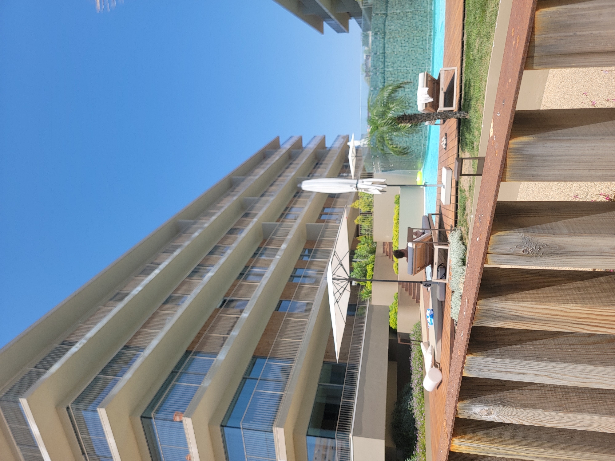 This media file depicts a sunny day scene at a resort or apartment complex in Quarteira, Portugal. In the foreground, wooden stairs with a rusty metal railing lead down to a poolside area. The poolside features a vibrant blue swimming pool surrounded by a wooden deck and lush green grass. There are lounge chairs and umbrellas by the pool, suggesting a relaxed atmosphere for sunbathing and swimming.
A modern, multi-story building with many balconies and large windows dominates the background. The architecture of the building appears contemporary, with clean lines and a light-colored facade. The windows reflect the bright blue sky, indicating clear weather. A palm tree is visible near the pool, adding to the tropical or Mediterranean feel of the location.
In the midground, a person is seated on a lounge chair near the pool, appearing to be reading or relaxing. Another individual's back is visible further down the stairs, suggesting movement within the area. A white, umbrella-like structure is also present, possibly a parasol or a decorative element.
The overall impression is one of a leisurely vacation setting on a clear, bright day. The sunlight casts distinct shadows, indicating it is likely mid-morning or afternoon. There is no visible text in the media file.