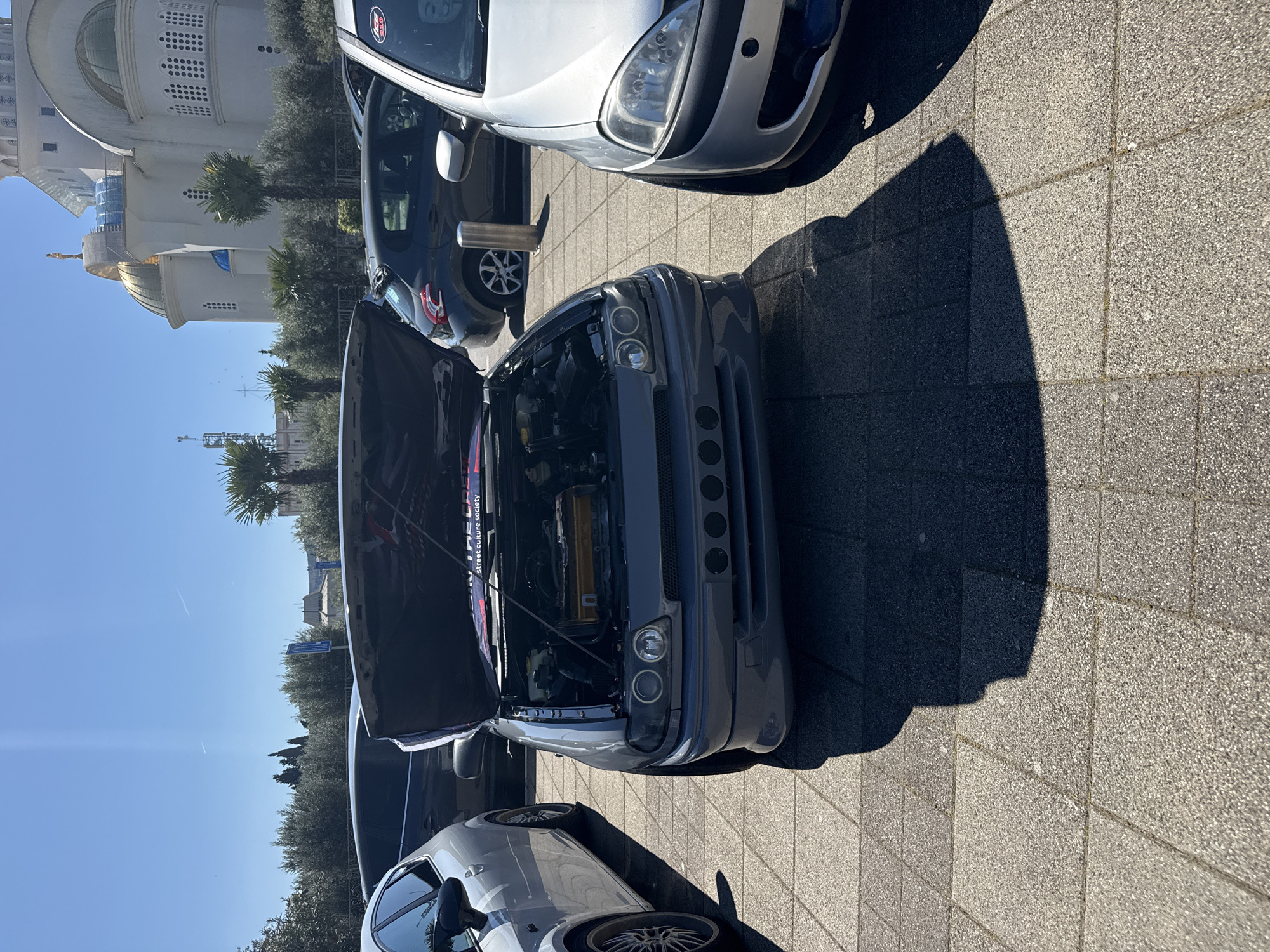 The image shows several cars parked on a paved surface. A gray car with its hood open is prominently featured, revealing the engine components. The backdrop includes buildings with architectural details such as domes and rows of windows. The image is taken outdoors in daylight, with clear blue skies visible. The scene appears to be located in a public area, possibly a parking lot, within the city of Unknown, Unknown.