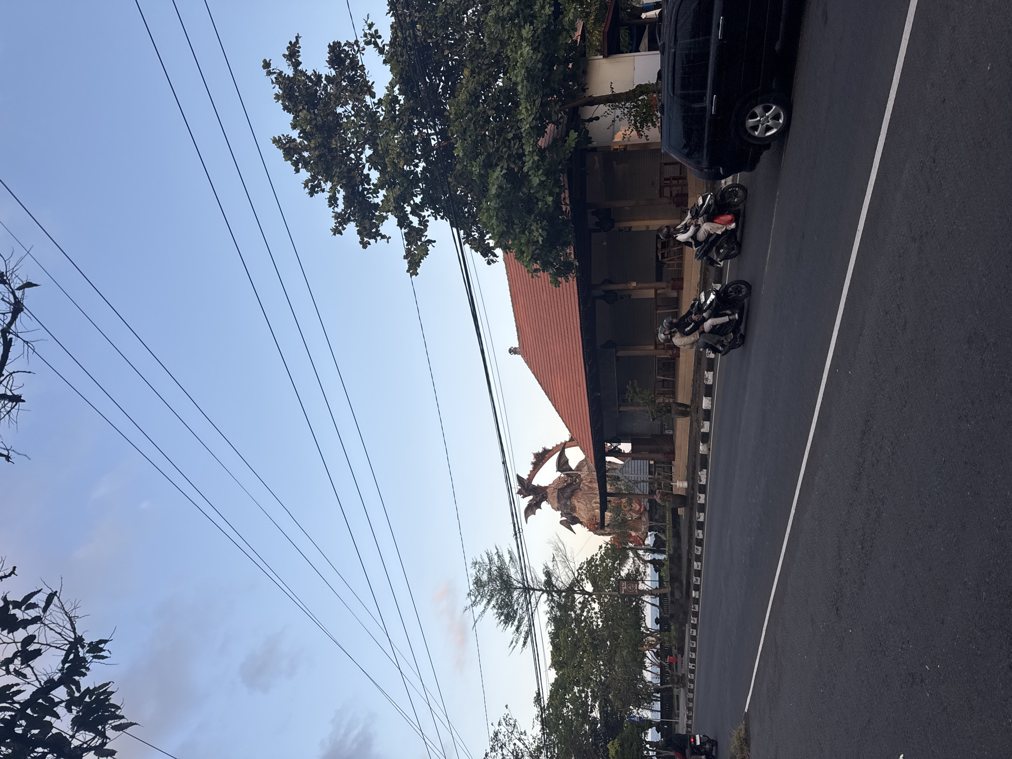 In Dawan, Indonesia, an overcast sky looms over a roadside scene. A dark-colored SUV is parked on the right side of a paved road, its passenger side wheel visible. Two motorcycles are traveling on the road in the same direction as the SUV. The first motorcycle carries two individuals wearing helmets. The second motorcycle is ridden by a single person, also wearing a helmet.
In the background, a building with a red tiled roof and an open-air veranda is situated behind a low wall. A large, imposing statue, appearing to be a mythological creature with wings and sharp features, stands behind the building. Trees are scattered throughout the scene, with some foliage in the foreground and more densely packed trees lining the area behind the wall. Electrical wires are strung across the sky, adding a linear element to the composition. The road itself is marked with a white line, dividing the lanes of traffic.