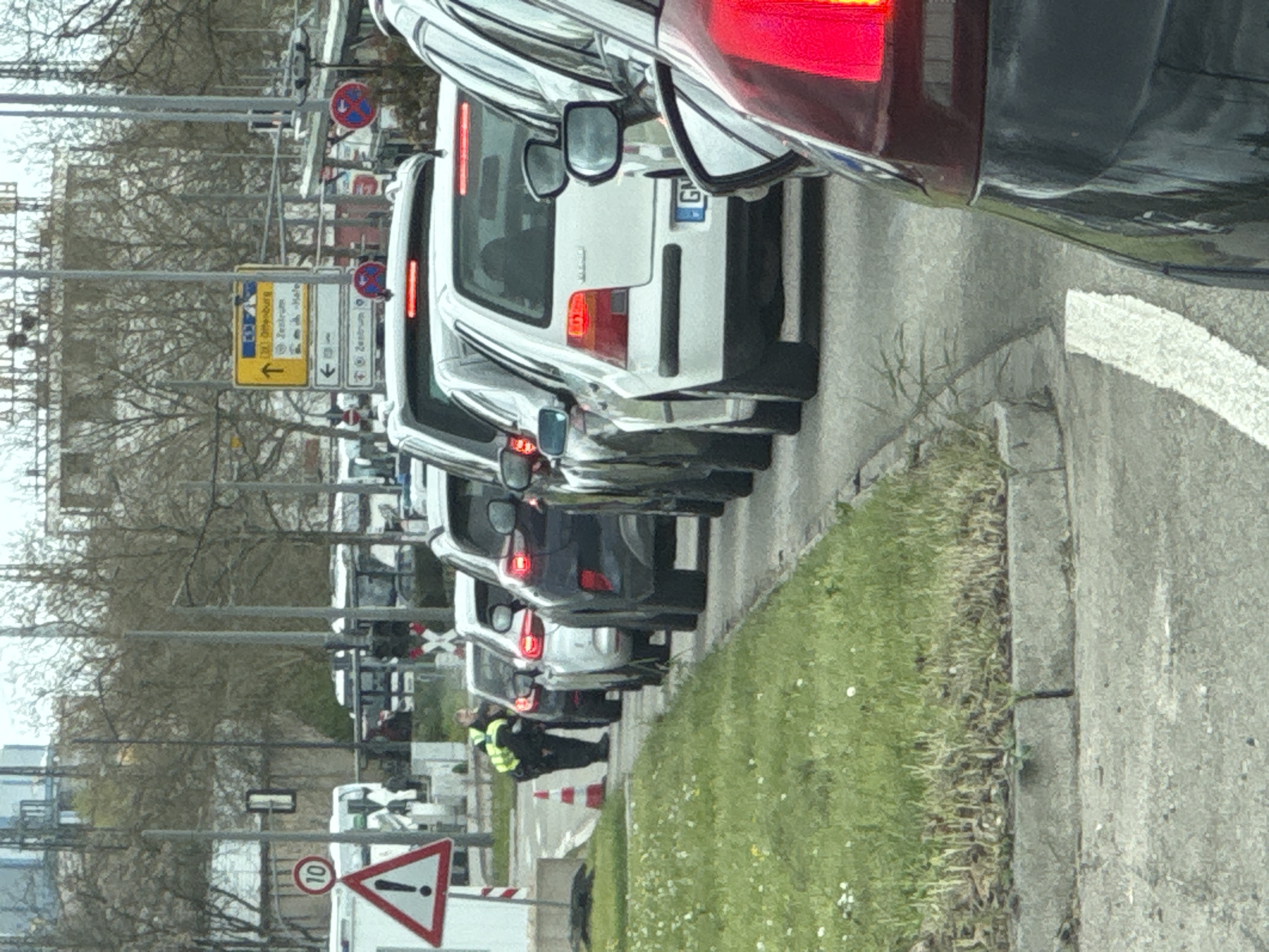 The image shows a line of cars stopped in traffic in Kehl, Germany. A person wearing a high-visibility vest is standing near traffic cones on the right side of the road. Road signs indicate the direction to "Zentrum" (center) and other locations. A speed limit sign with a "10" is visible. The license plate of the second car in line begins with "G".