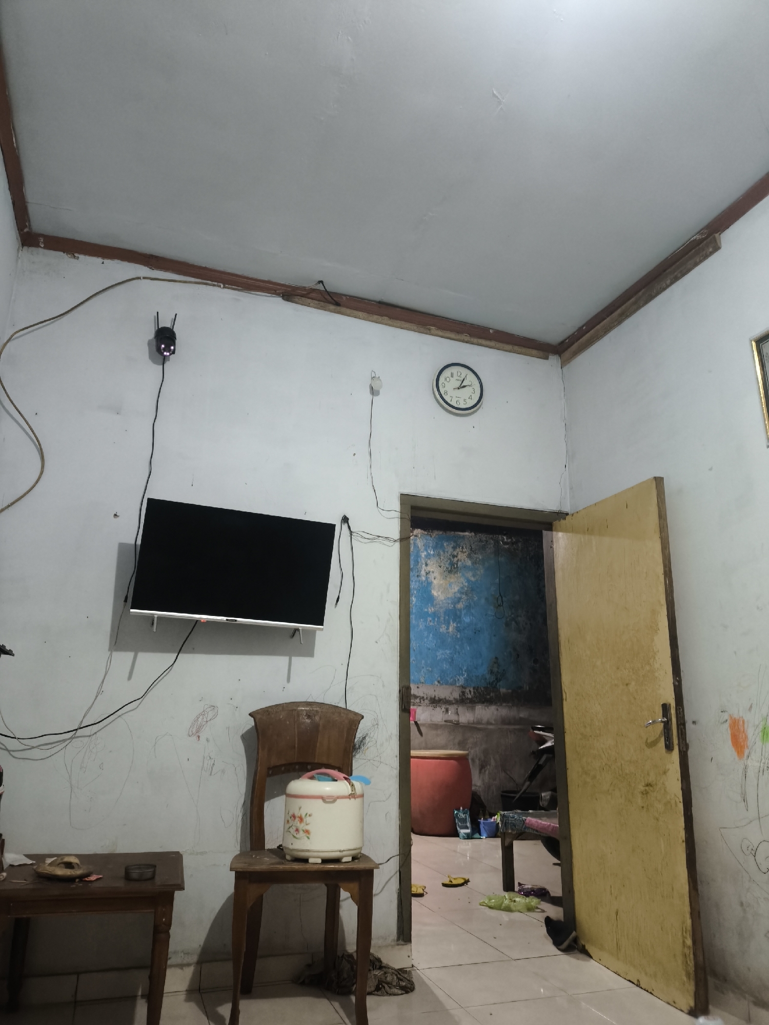 The image depicts a dimly lit interior room, likely a living space, with elements suggesting it is located in Senduro, Indonesia. The walls are painted white and appear somewhat aged, with visible cracks and what might be child-like drawings or scribbles. The ceiling is also white, with wooden trim along the edges where it meets the walls.
In the center-left of the frame, a flat-screen television is mounted on the wall. Above the television, a small black device, possibly a Wi-Fi router or a security camera, is attached to the wall. To the right of the television, a vintage wooden chair is placed, and on the seat of the chair sits a white rice cooker adorned with a floral pattern.
In the foreground, to the left, a small wooden table holds a pair of flip-flops and a small circular object, possibly an ashtray or a container.
To the right, an open wooden door reveals a glimpse into another area, which appears to be a darker, possibly utilitarian space. The wall in this secondary area is painted a distressed blue and gray. A large red cylindrical object is visible in this space, along with what looks like a motorcycle.
A round wall clock with a white face and black numerals is mounted on the wall above the doorway, indicating the time is approximately 12:08. The floor appears to be tiled. The overall impression is of a lived-in, modest dwelling, with everyday objects and signs of recent use.
