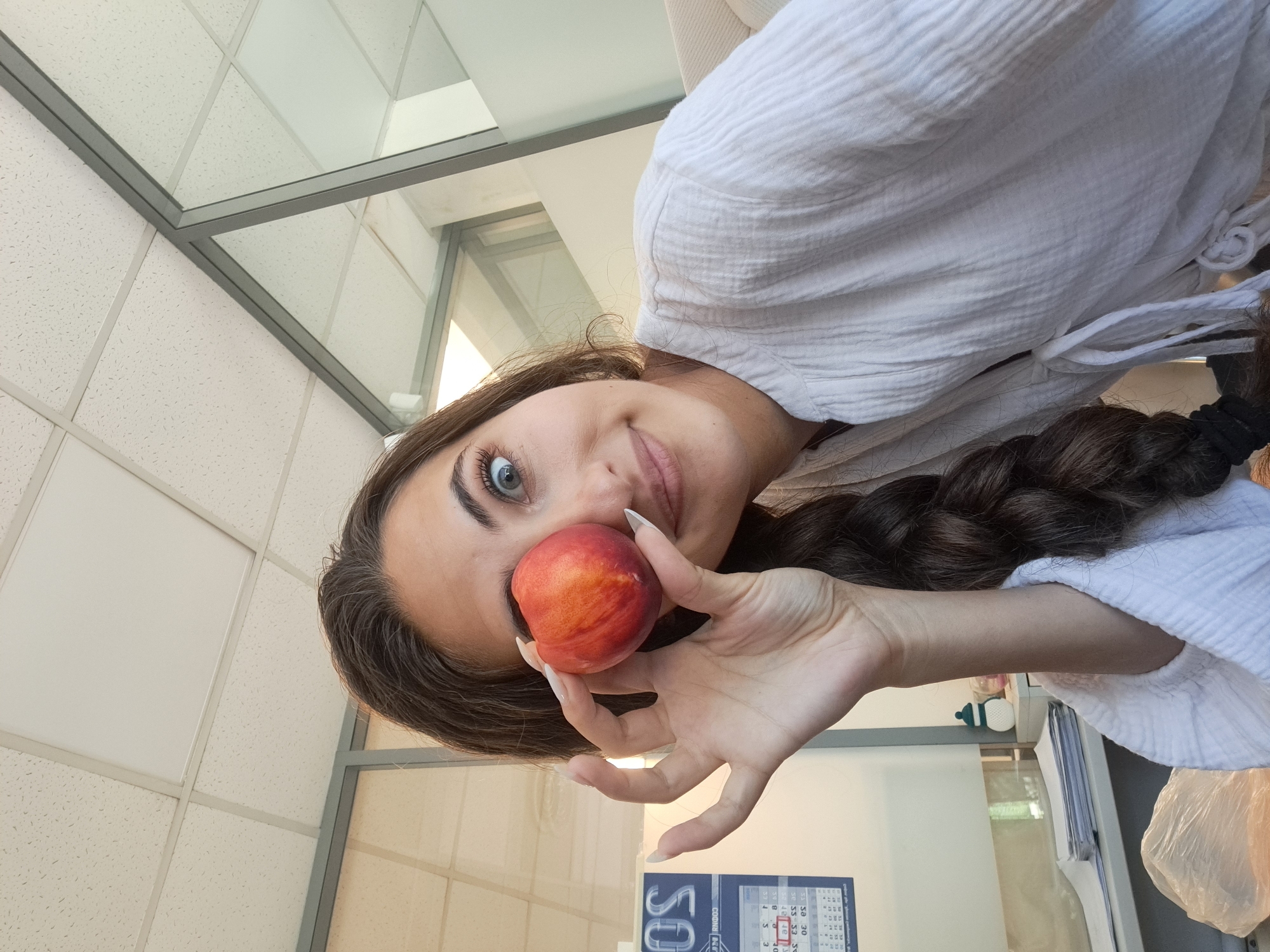 A young woman with long brown hair braided down her back is holding a nectarine up to her left eye, covering it, and looking at the camera with a surprised expression. She is wearing a white gauze-like shirt. The background appears to be an office environment with a tiled ceiling, glass partition walls, a desk with papers, and a calendar hanging on the wall. The calendar shows the month of August 2024, indicating the possible location in Sofia, Bulgaria, as indicated by the text "SOFIA" visible on the calendar. The lighting suggests an indoor setting, possibly during daytime.