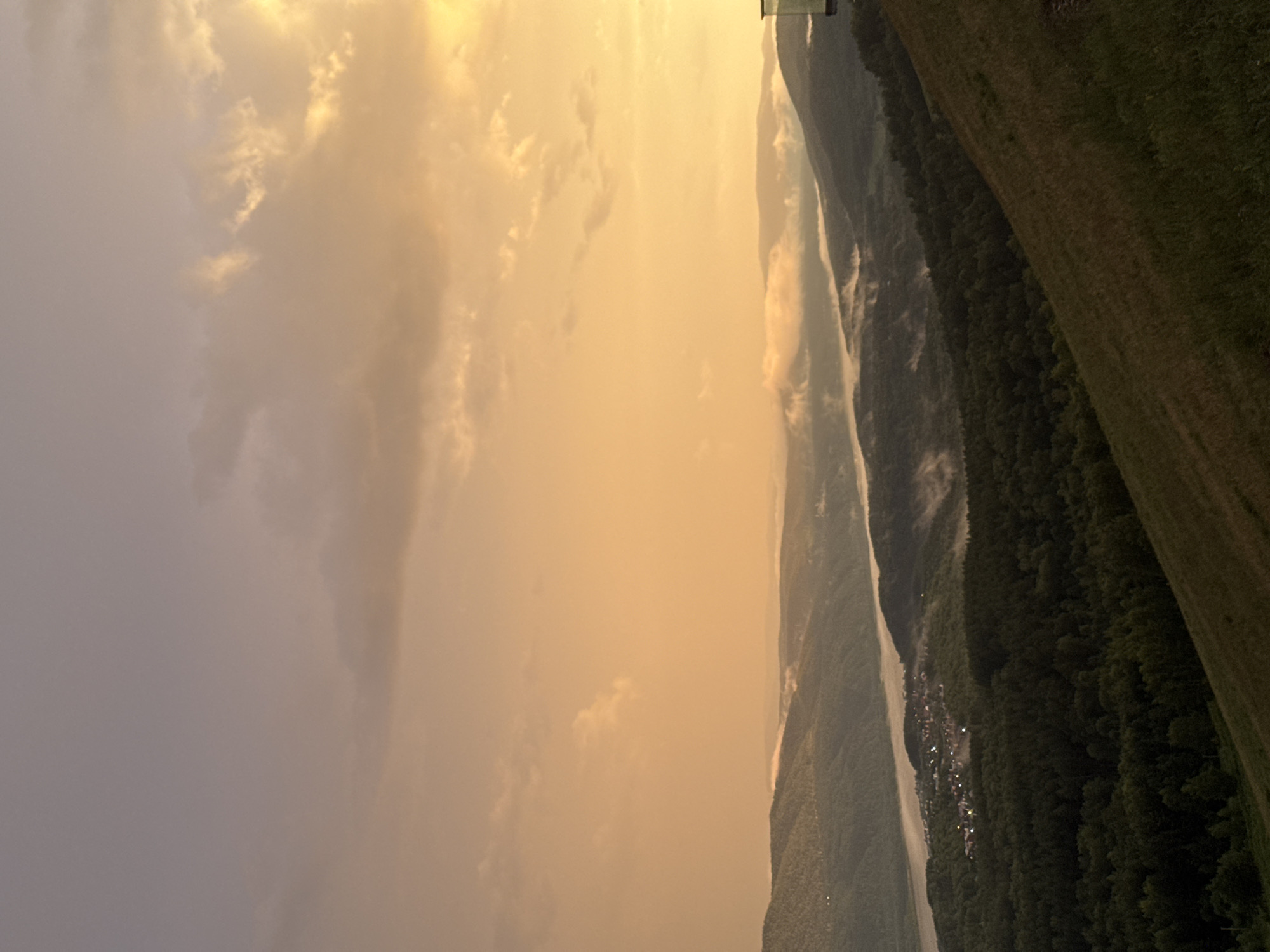 This media file shows a landscape scene captured during what appears to be sunset or sunrise, given the warm, golden hues of the sky and the soft, diffused light. The sky is filled with scattered clouds, some catching the light and appearing bright yellow, while others are darker and more diffused, creating a layered effect.
The dominant feature of the scene is a vast valley with rolling hills and mountains. A river or large body of water snakes through the valley, reflecting the sky's colors. The hillsides are densely covered in dark green forest. In the distance, nestled within the valley, a small settlement with scattered lights can be observed, suggesting a human presence. The light is low, casting long shadows and contributing to a serene, tranquil atmosphere. The overall mood is one of natural beauty and peacefulness. There is no visible text in the image. The location context provided is Krasnoyarsk, Russia.