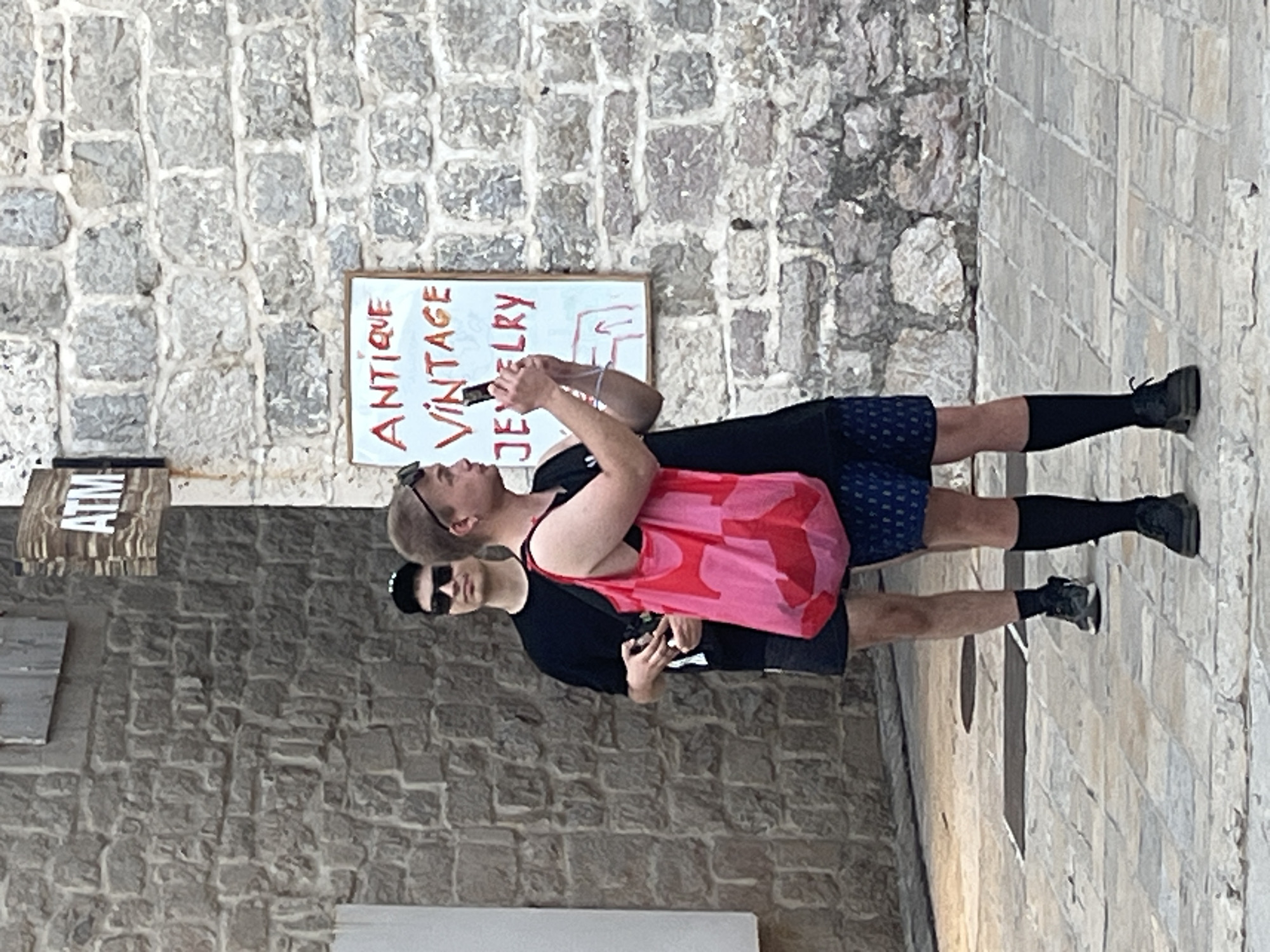 Two individuals stand on a cobblestone street in front of a stone building. The person in the foreground, facing right, is wearing black socks, black shoes, blue patterned shorts, and a black t-shirt. They are holding a smartphone in their right hand, appearing to take a photo. A bright pink tote bag with a red pattern is slung over their left shoulder. The second person, partially visible behind and to the left of the first, wears black shorts, black socks, black shoes, and a black t-shirt with a black baseball cap and sunglasses. They are holding a camera or phone in their hands. A white sign with red lettering reads "ANTIQUE VINTAGE JEWELRY" on a wooden frame. An "ATM" sign is visible above and to the left of the jewelry sign. The architecture suggests an old town setting, consistent with the location of Budva, Montenegro.
